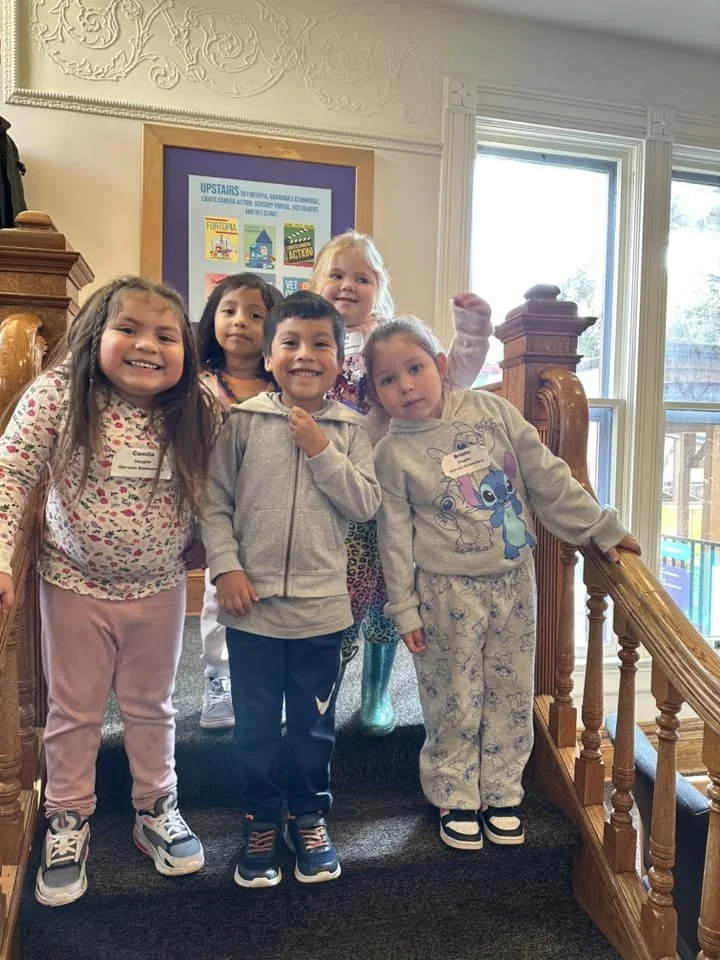 Five smiling children standing on the historic wooden staircase inside the museum. An "Upstairs" exhibit sign is visible on the wall behind them.