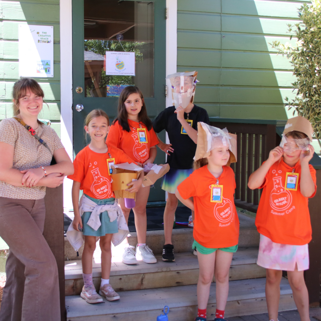 Group of children and an adult standing on a porch during a summer camp activity. Some children are wearing paper hats and goggles, holding containers, with a green house exterior in the background.