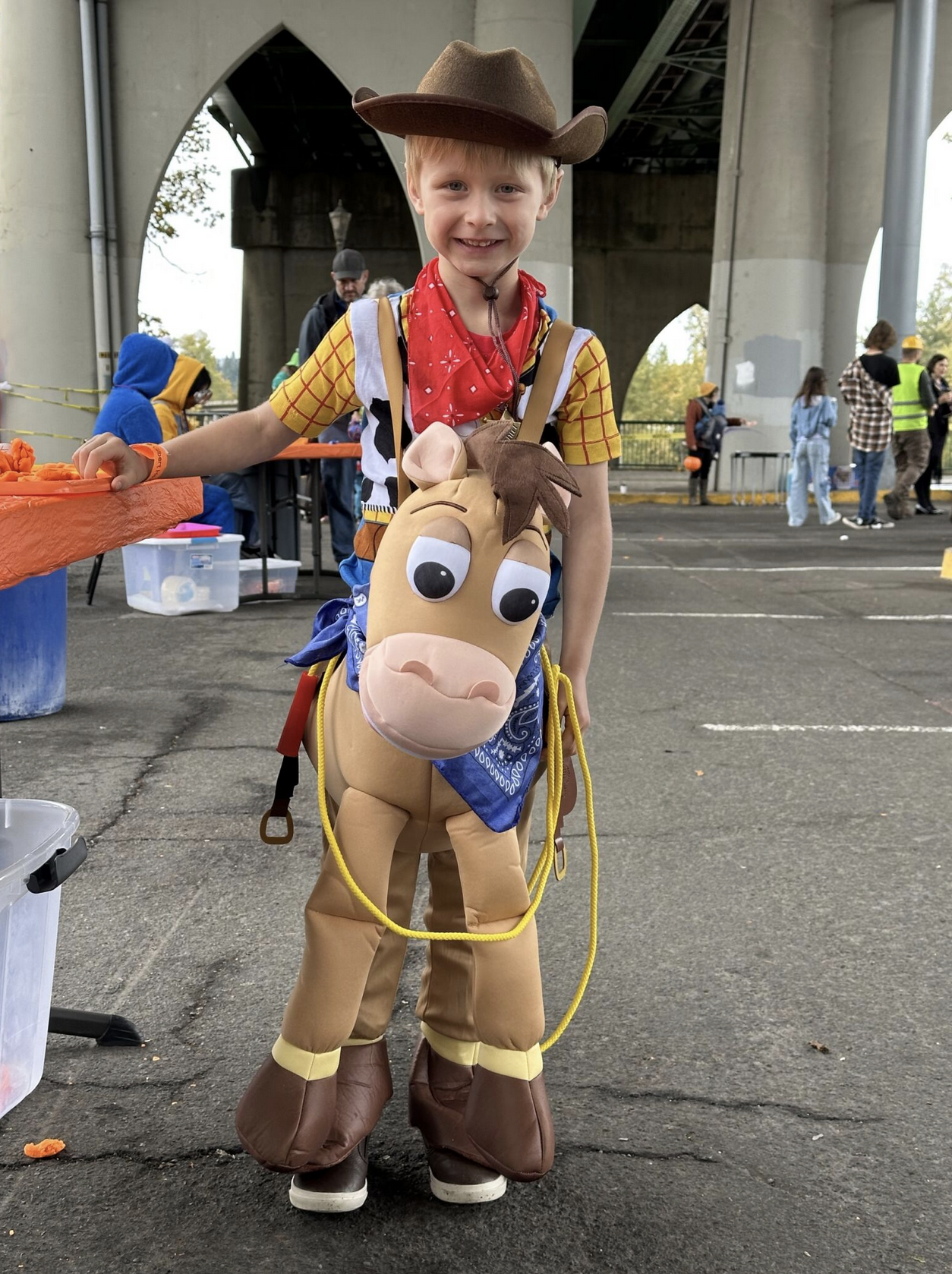 A young boy dressed as Woody from Toy Story, wearing a cowboy hat and a toy horse costume, smiling at the camera under a bridge at an outdoor event.