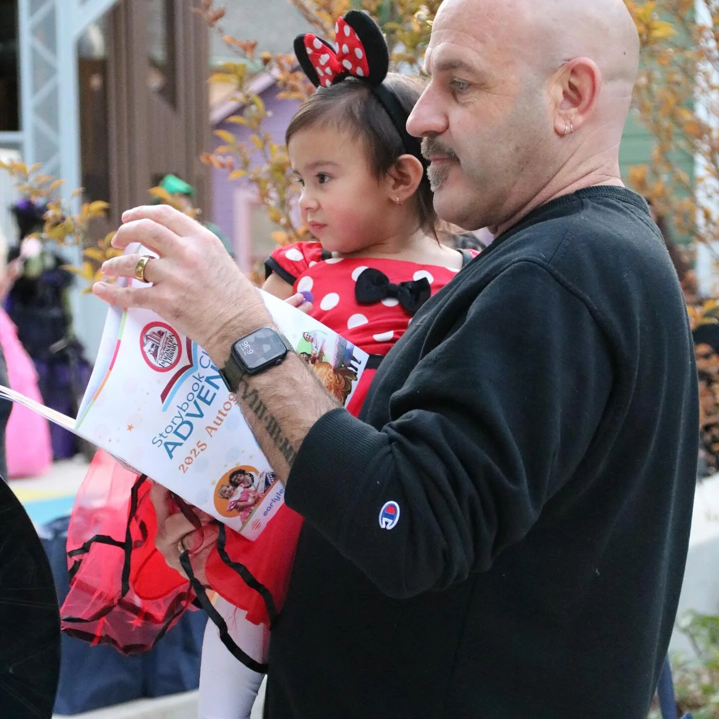 A man shows a Storybook Adventure guidebook to a toddler in a Minnie Mouse costume at a private family event.