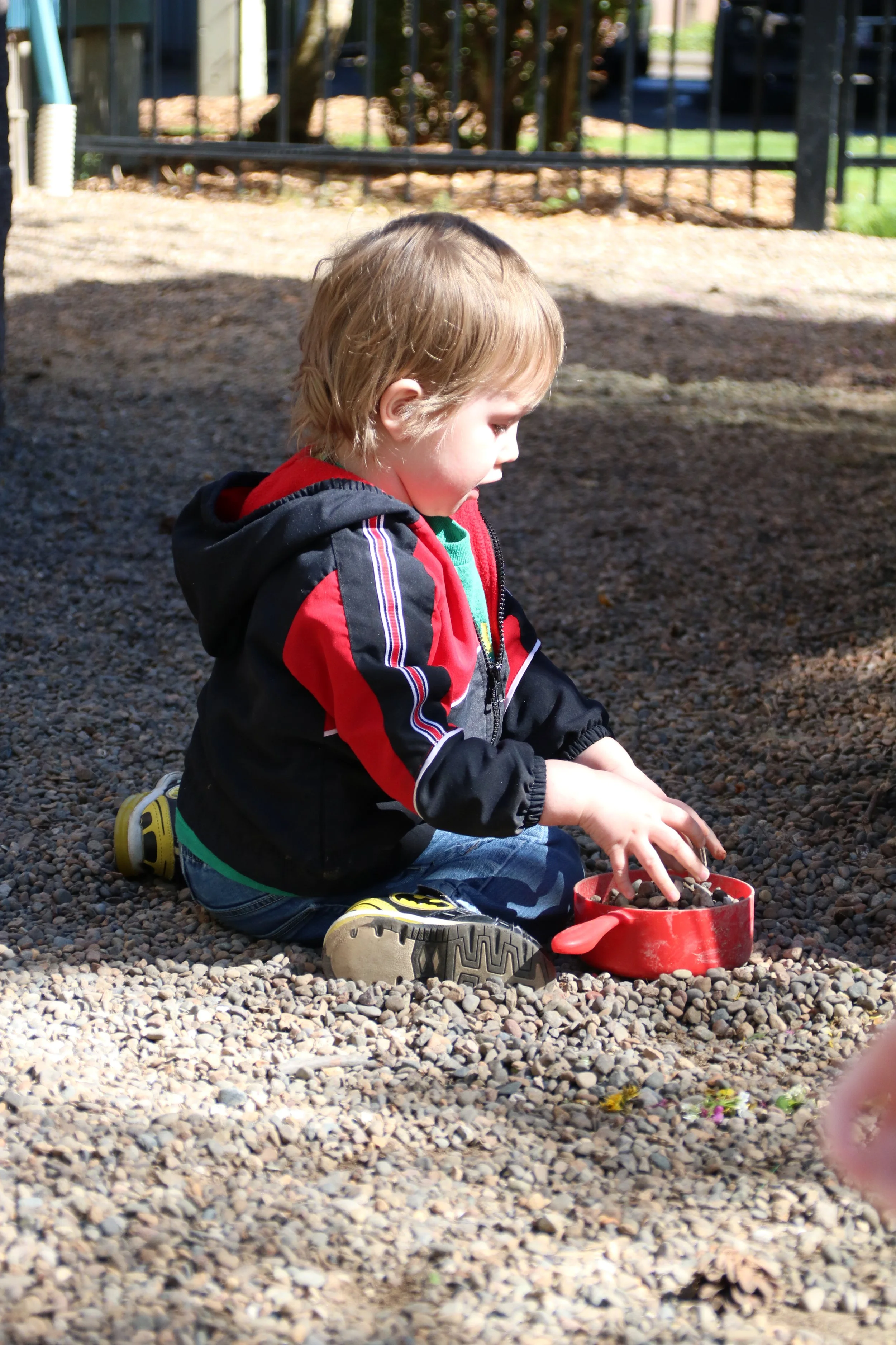 A young child with blonde hair, wearing a black and red jacket, kneeling on a gravel surface outdoors, playing with a red toy shovel and small rocks.
