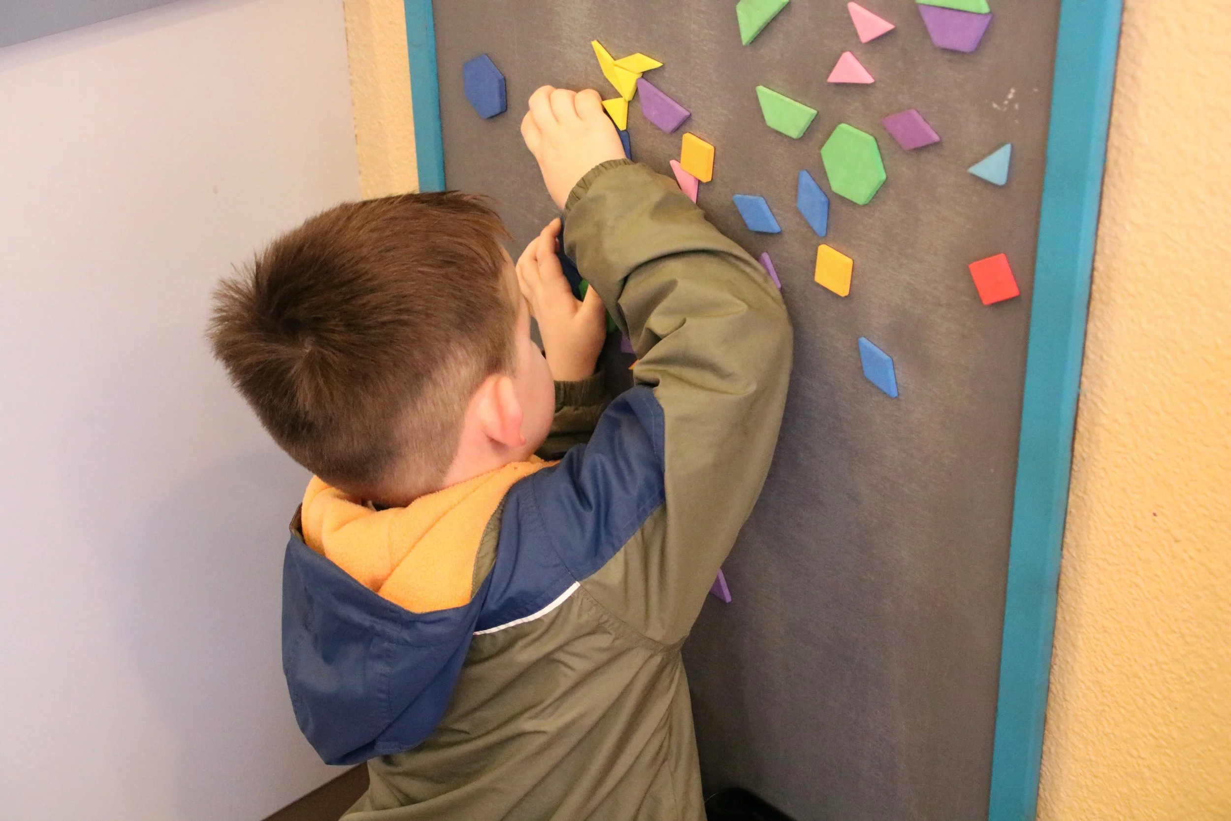 A young boy with short brown hair wearing a tan, blue, and yellow jacket arranging colorful wooden geometric shapes on a black felt board.