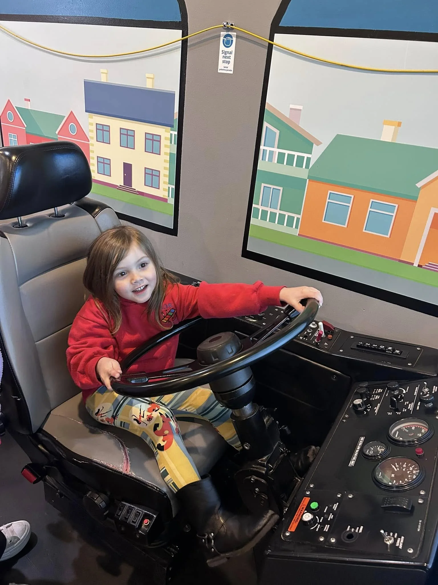 Young girl sitting in a bus driver's seat holding the steering wheel, smiling. Behind her are colorful posters of houses on the wall.