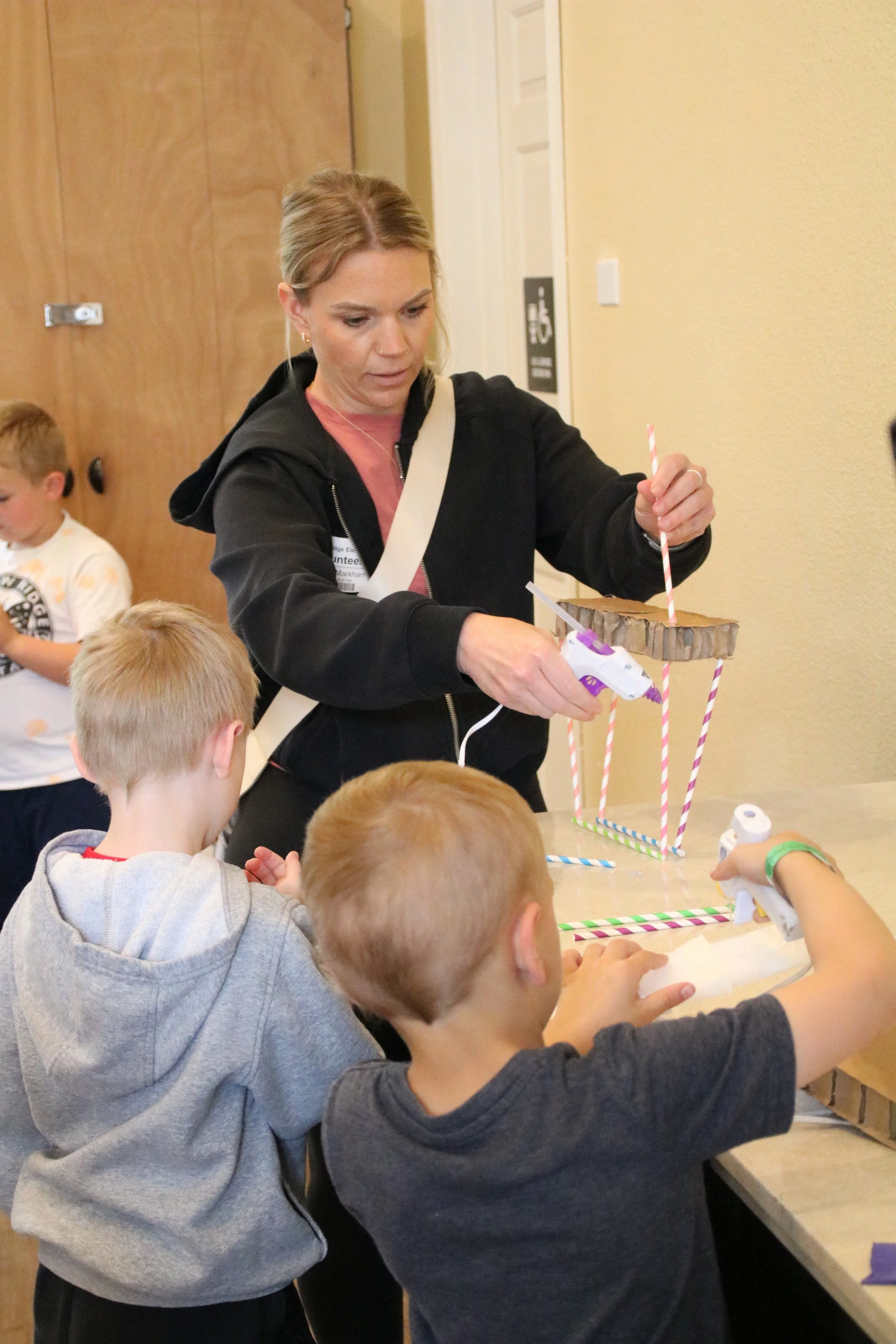Woman and children engaged in arts and crafts activity at a table, creating a structure with sticks, glue, and cardboard.