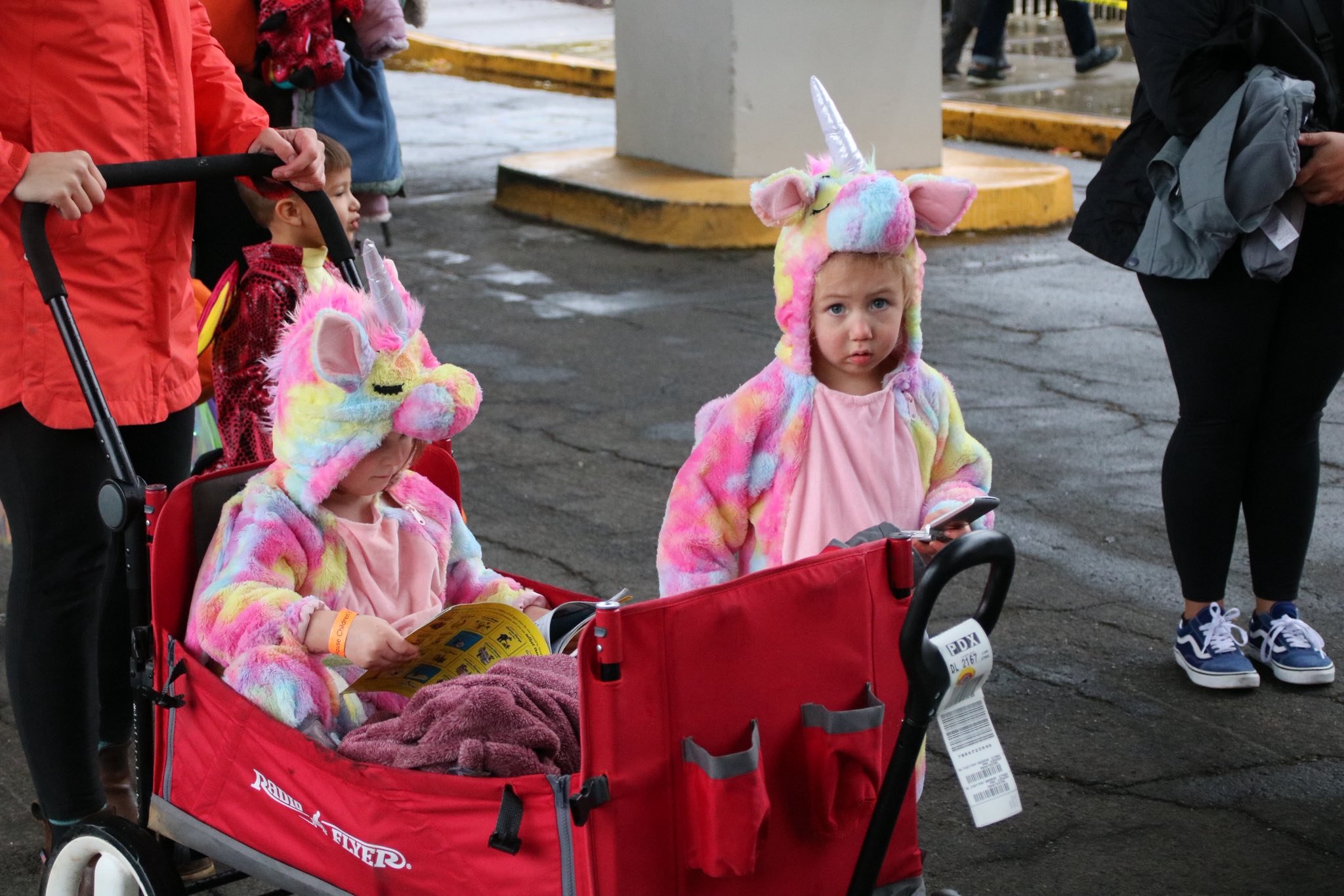 Two young children dressed in colorful unicorn costumes, one sitting in a red wagon reading a brochure and the other standing next to it, holding a phone, with adults around them on a street in rainy weather.