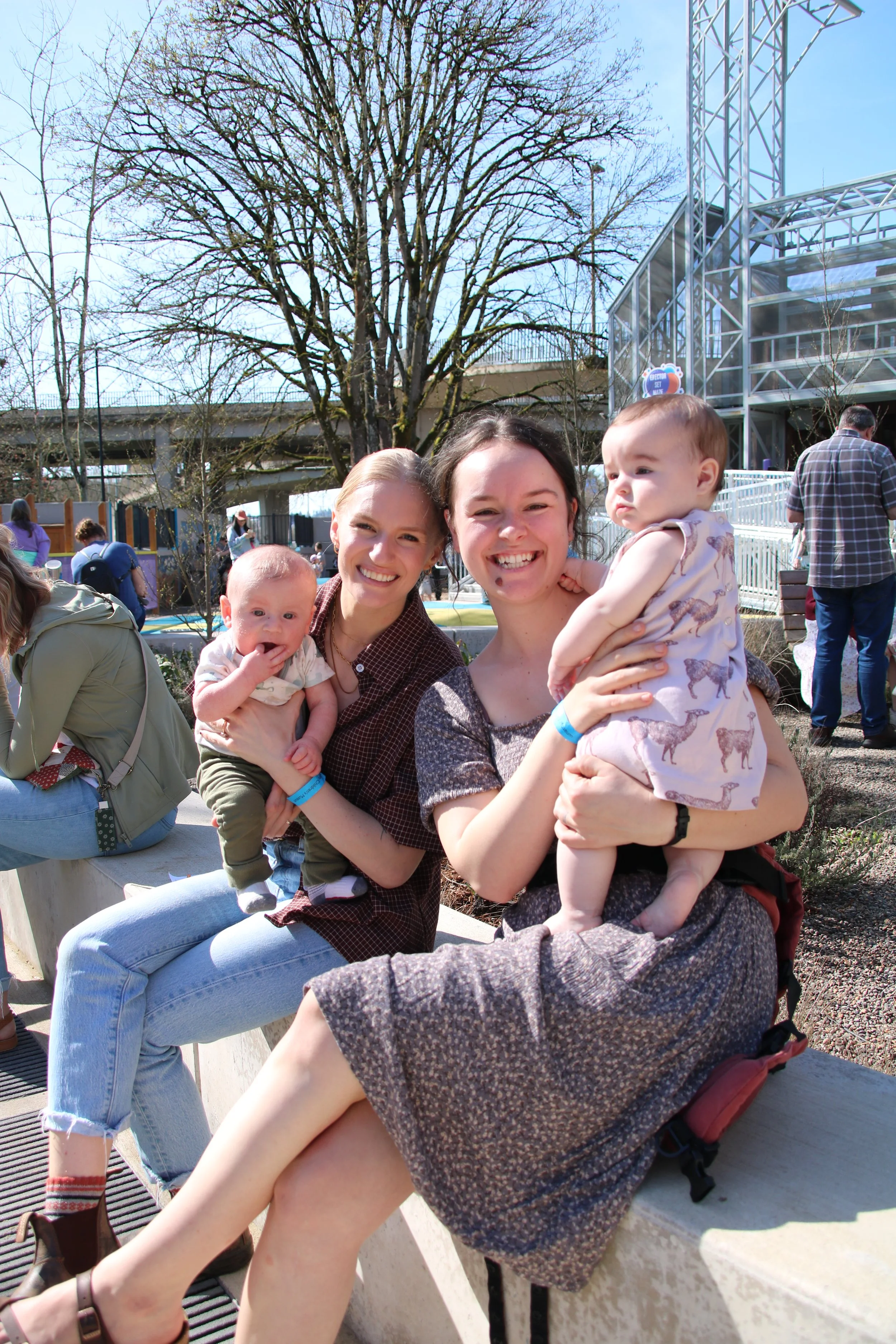 Three women with two children sitting on a concrete bench outdoors at an amusement park, with some people and rides visible in the background.