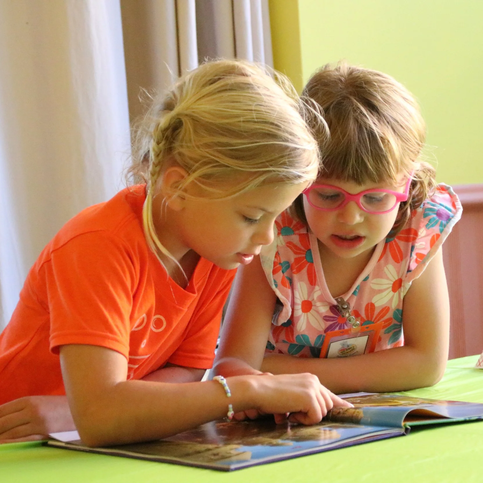 Two young girls reading a picture book together, leaning over the table, with one girl wearing pink glasses and a floral dress, and the other girl in an orange shirt, in a brightly lit room.