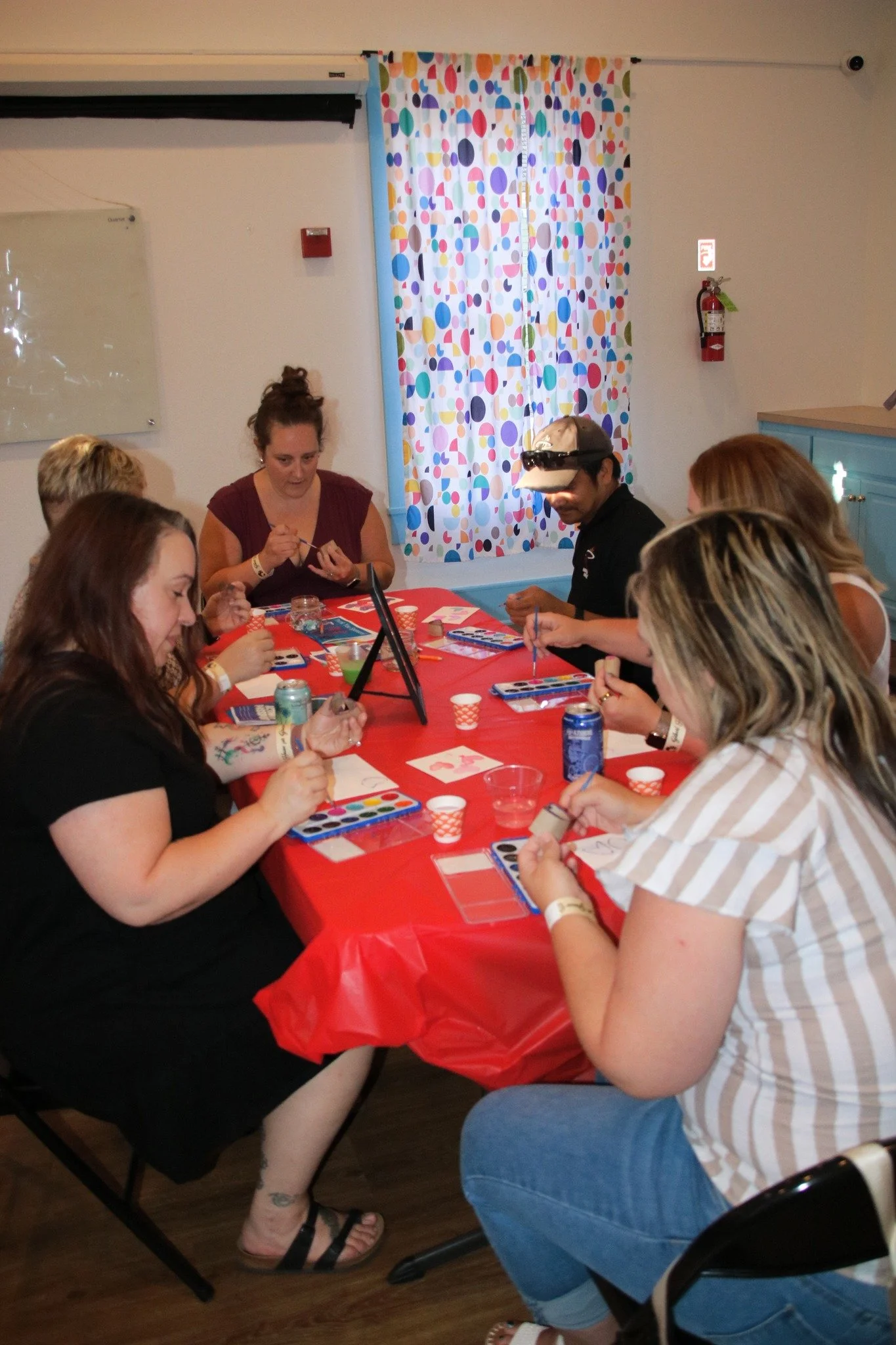 Group of women and one man sitting around a red table engaging in arts and crafts activities at a party. The man is wearing a headset and glasses. The room has a colorful polka dot curtain and a blue cabinet.