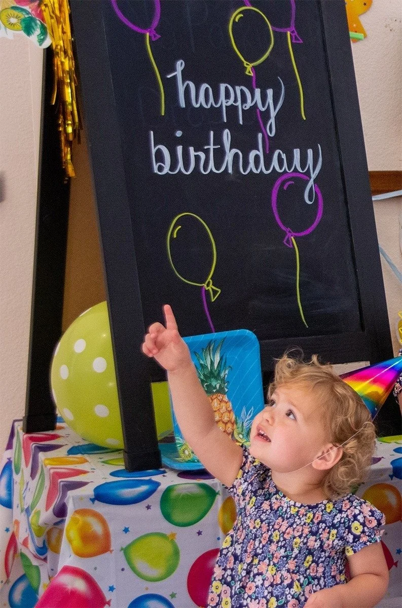 A young girl in a pink flower print dress and a rainbow striped party hat points excitedly. A "Happy Birthday" sign and a balloon-print tablecloth are visible in the background.
