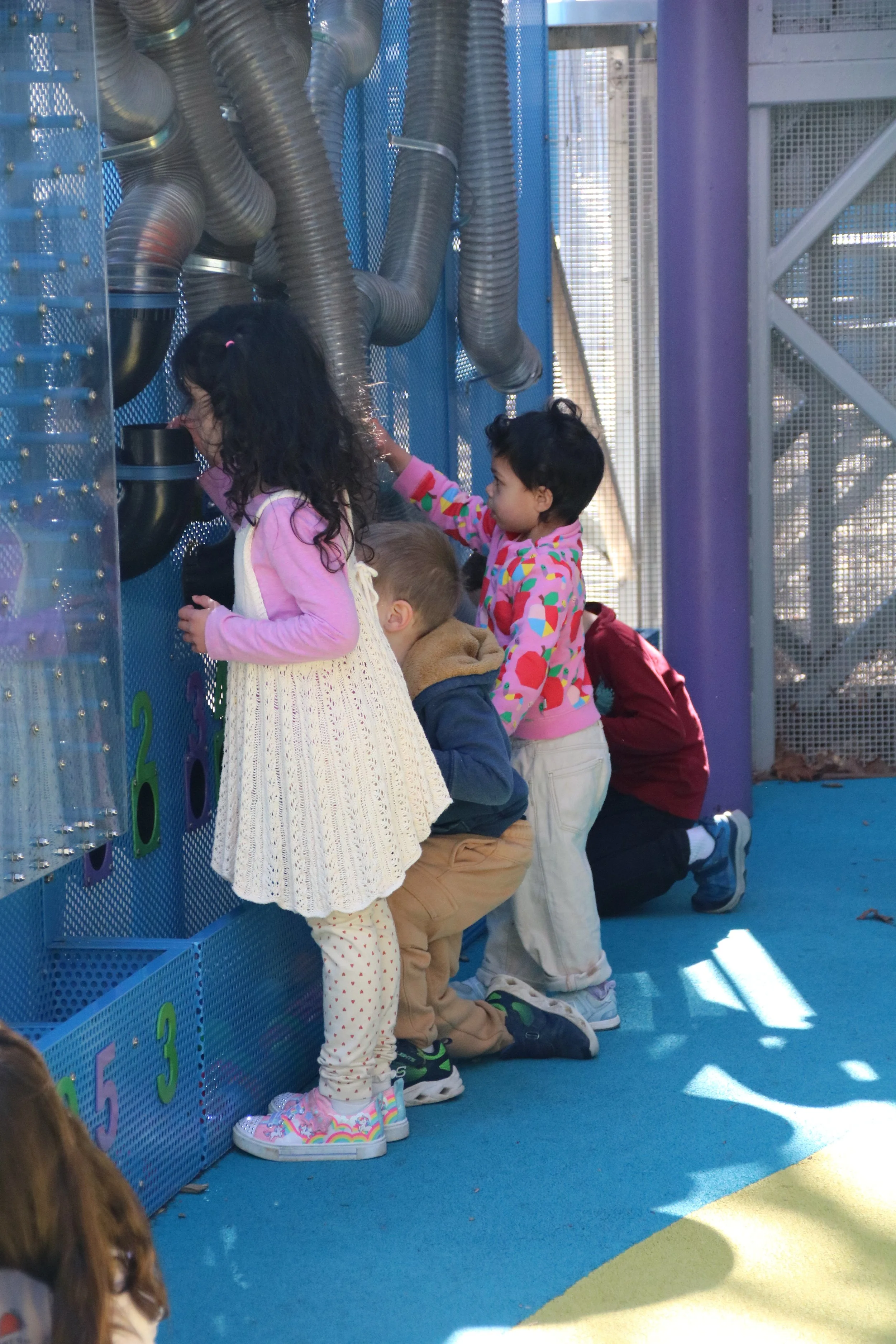 Children kneeling and standing in line, looking at a blue wall with colorful numbers and metallic tubes, at an outdoor playground.