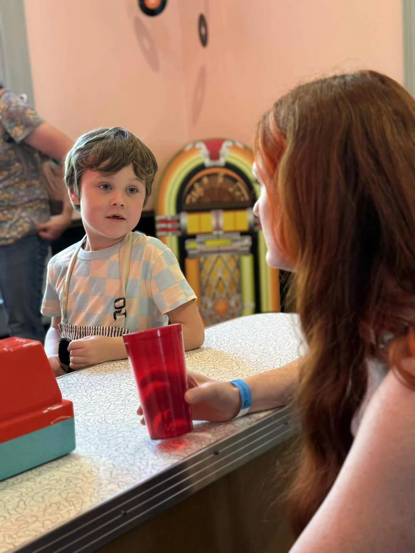 A young boy with short, brown hair wearing a light checked shirt and striped apron, talking to a woman with long red hair. The woman is holding a red cup and wearing a blue wristband. There is a vintage jukebox in the background.