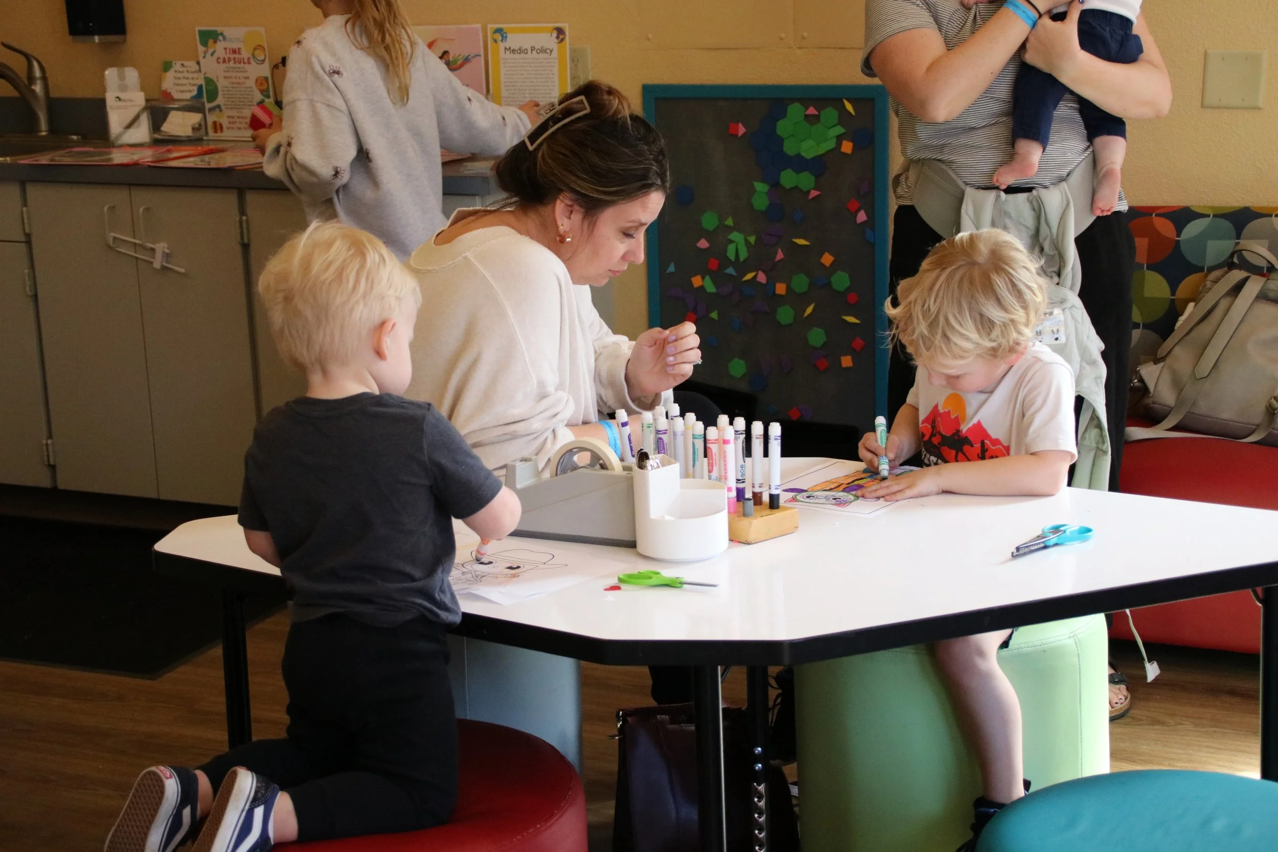 Children drawing with markers at a table while adults observe in a room with educational decorations.