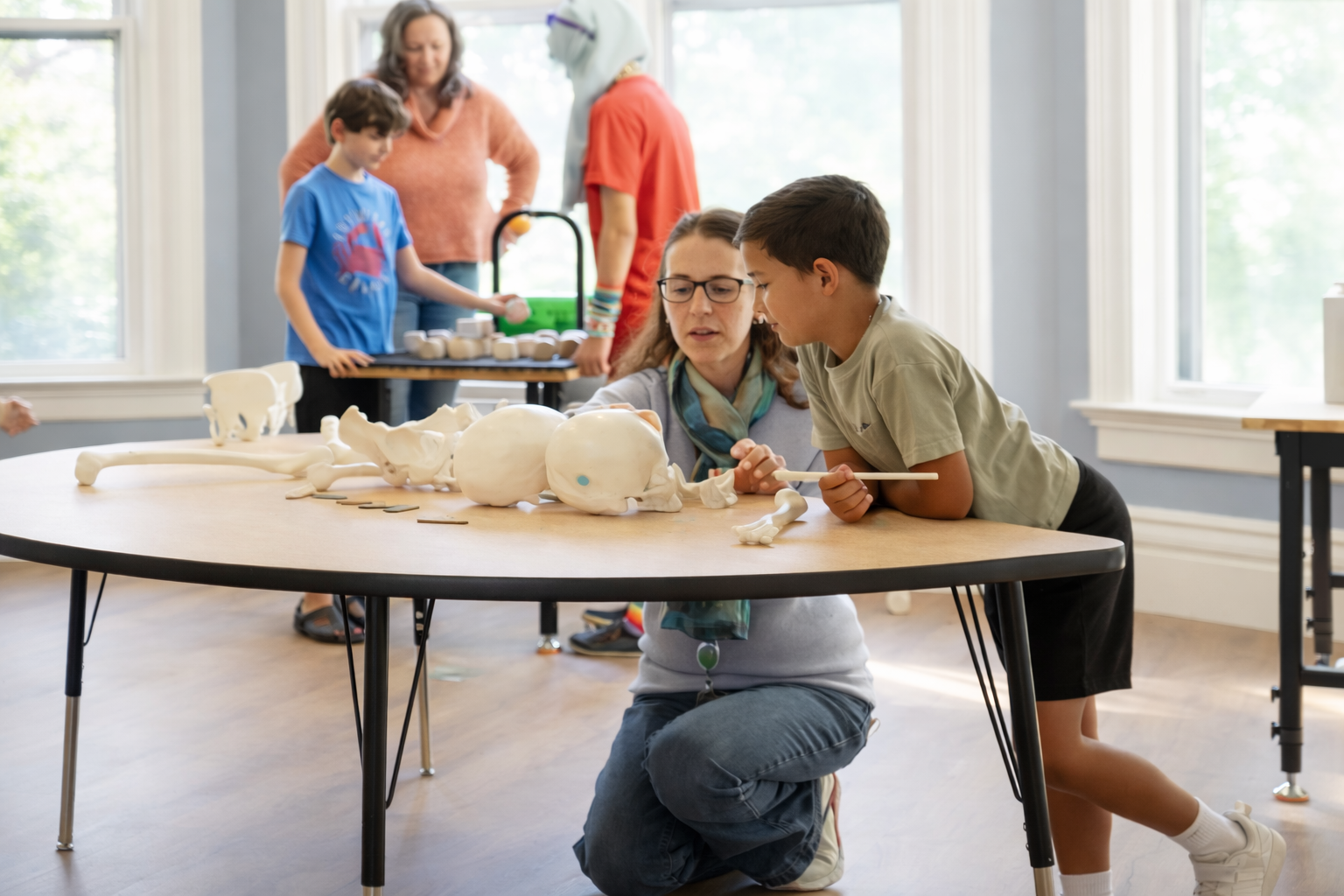 Children and adults around a table working on human skeleton models and bone replicas during an educational activity in a bright room.
