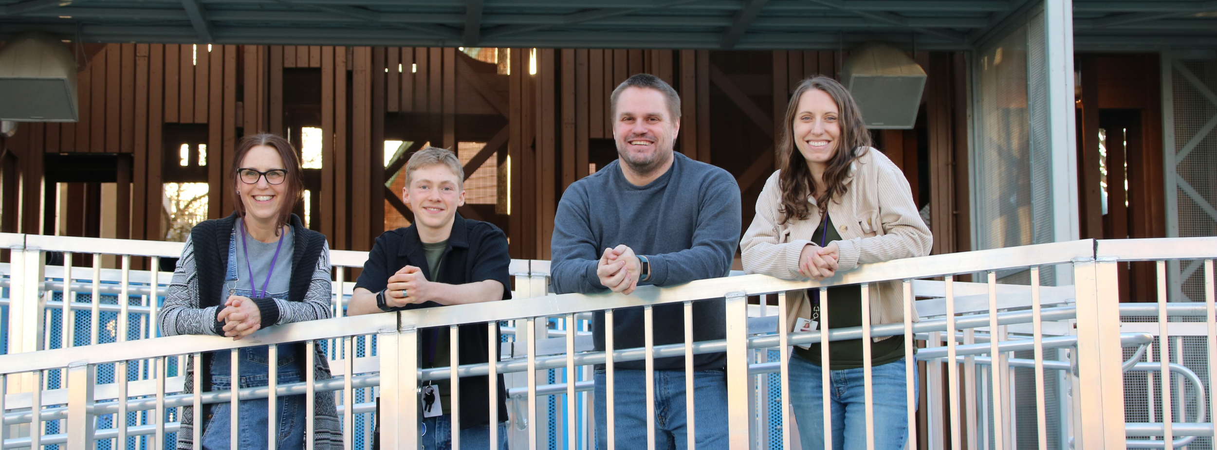 Four people smiling and leaning on a white railing outdoors, with a wooden building structure in the background.