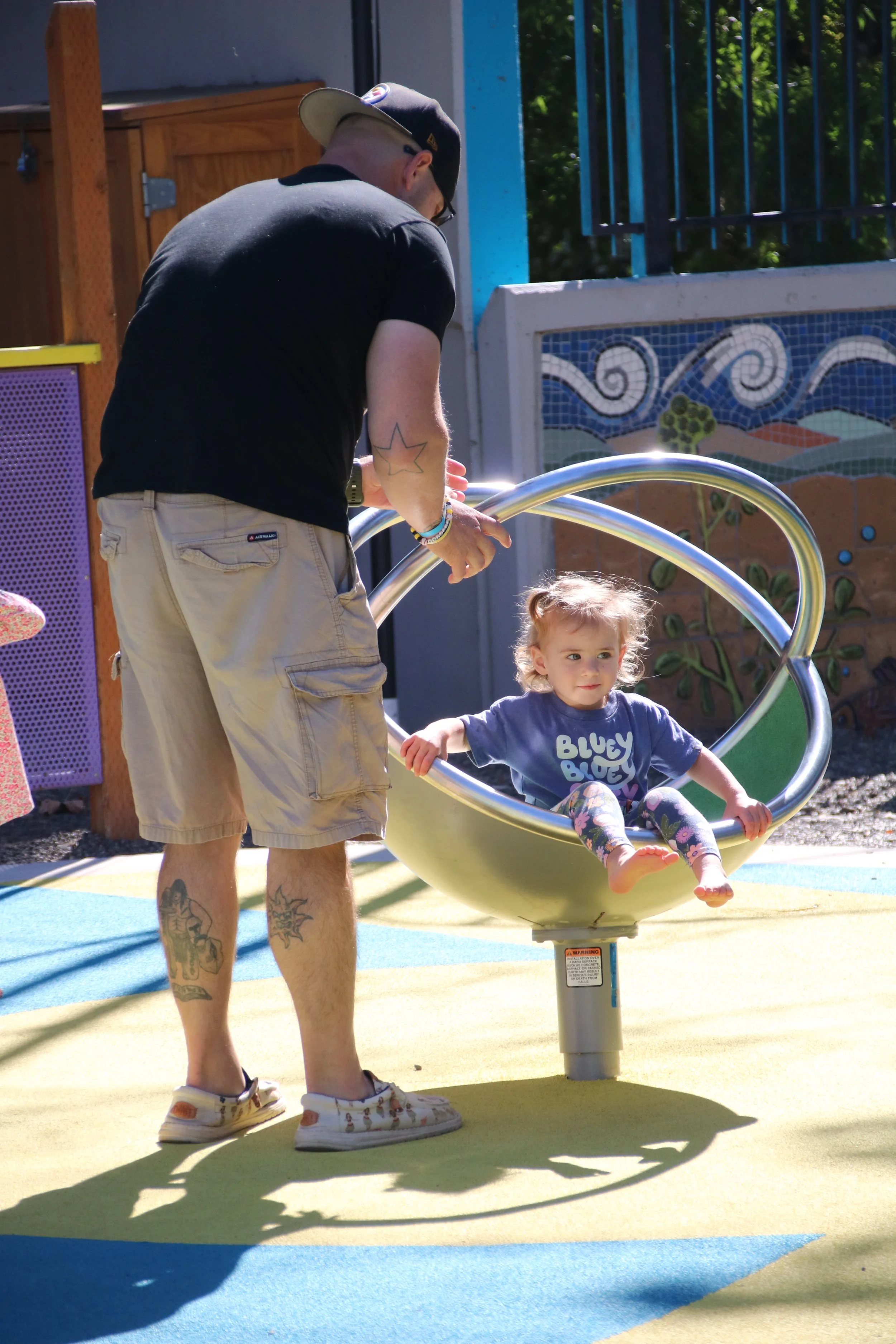 A man pushes a young girl in a yellow circular swing at a playground on a sunny day.