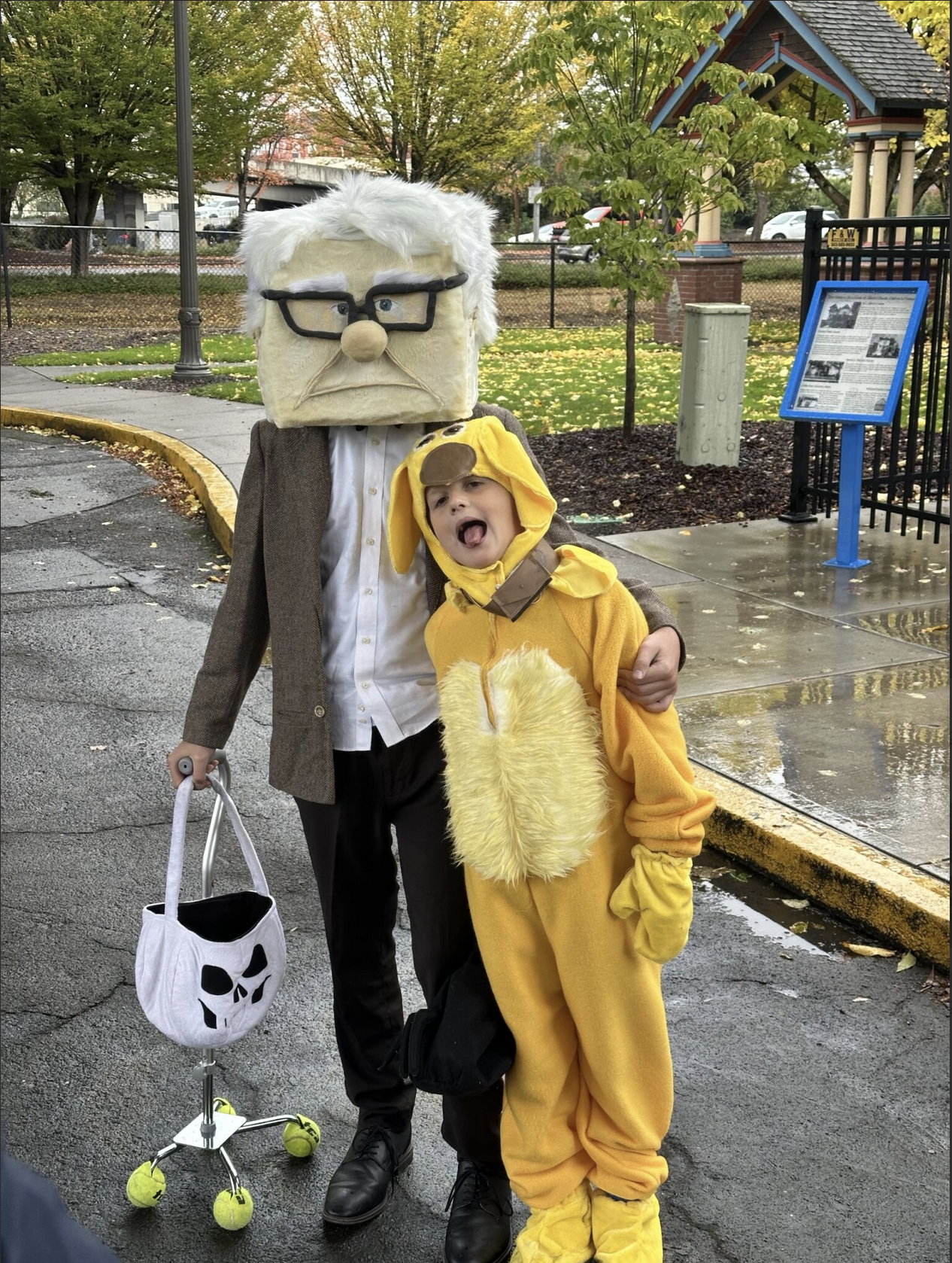 A person dressed as an elderly man with a large square head costume with white hair, glasses, and a mustache, standing outdoors on a wet pavement. They are holding a small white bag with a skull design on it. A young child in a bright yellow dog costume with floppy ears and a furry tummy is standing beside them, sticking out their tongue and making a funny face. The background shows autumn trees, a park, and a blue information sign.