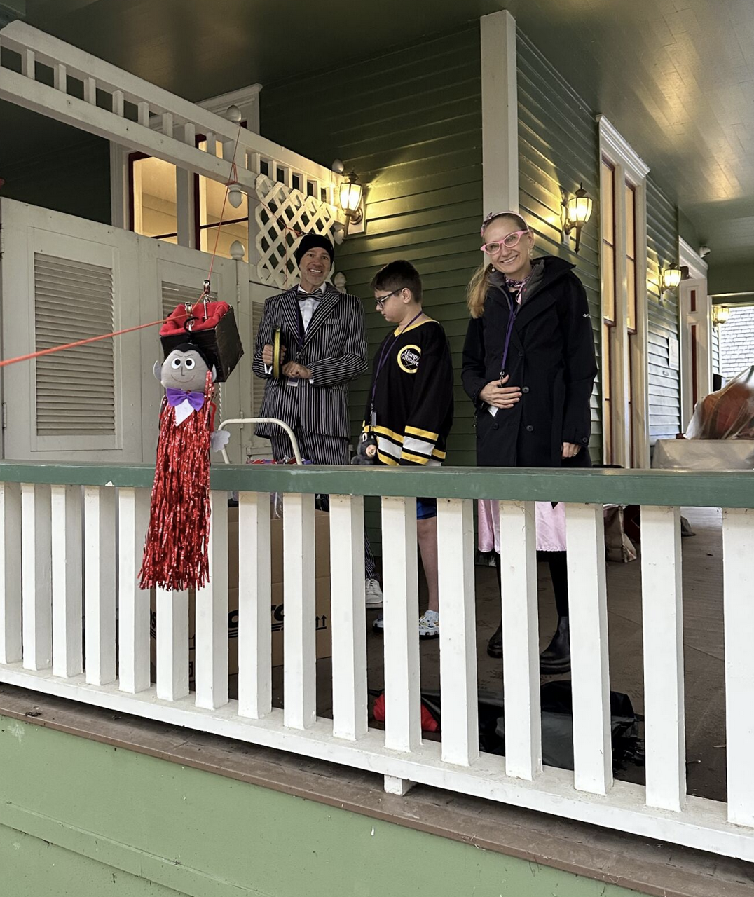 Three people dressed in costumes standing on a porch with a puppet hanging on a string in front of them.