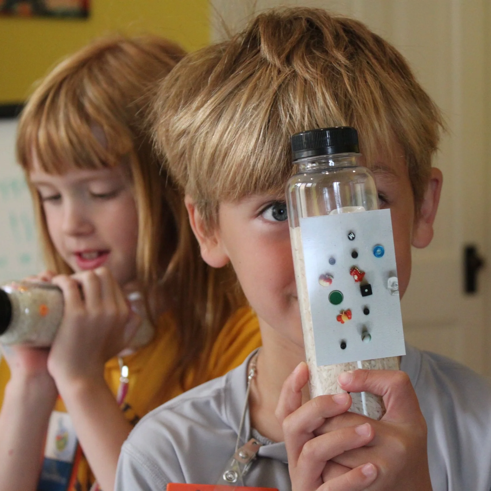 Two children, a girl with red hair and a boy with light brown hair, are conducting a science experiment with homemade binoculars containing beads and small objects inside.