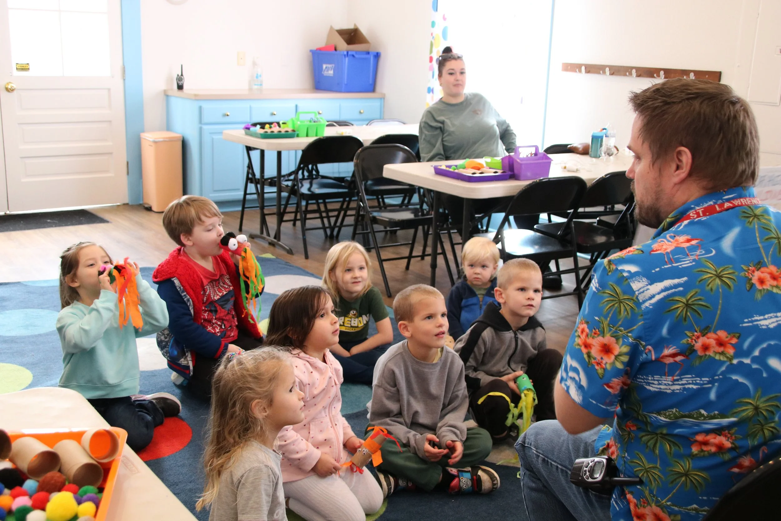 A group of young children sitting on a colorful rug, attentively watching a man who is talking to them. The children are holding crafts or toys, and the setting appears to be a classroom or activity room with tables and chairs in the background.