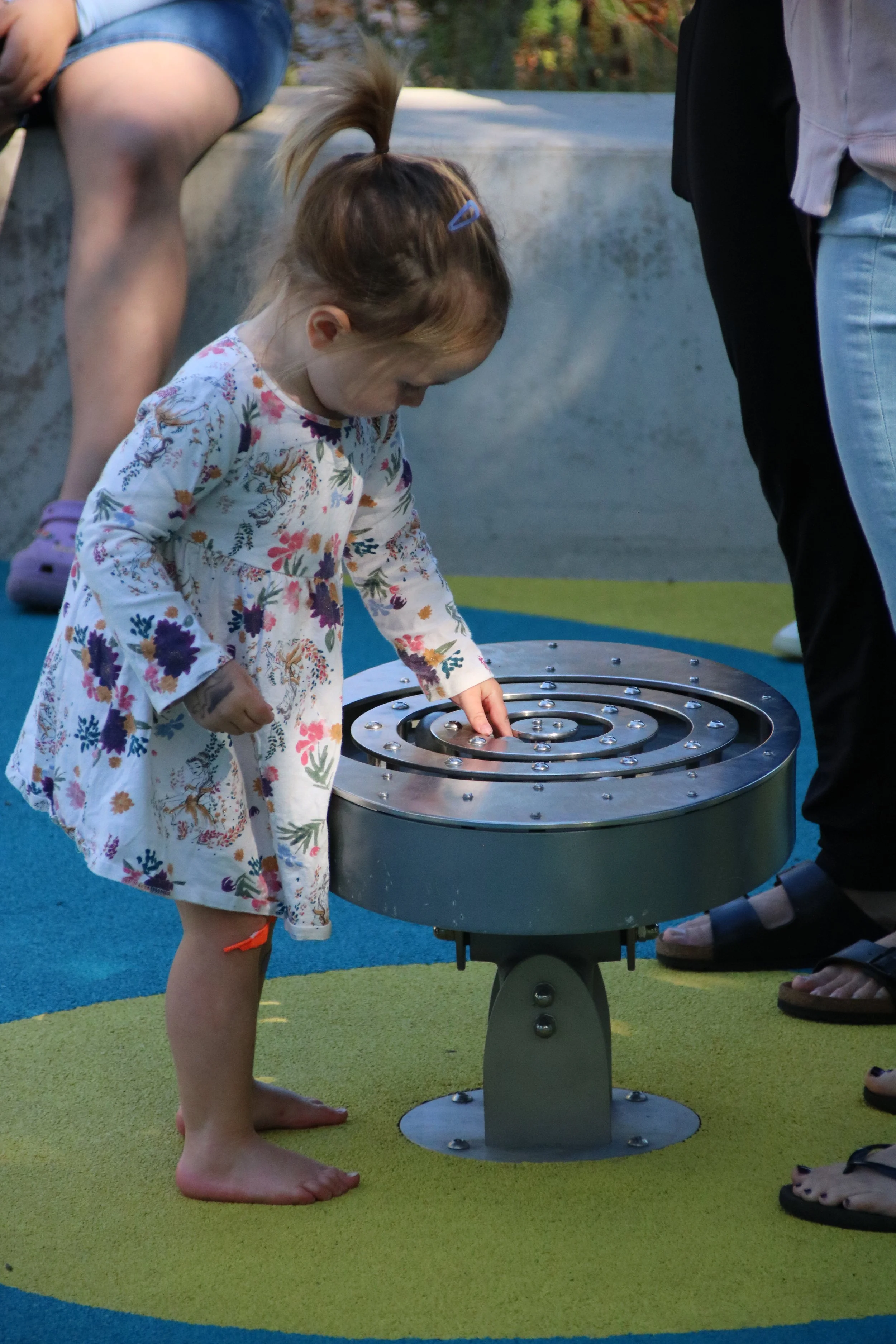 A young girl in a floral dress playing with a metal spiral-shaped musical instrument on colorful playground flooring, surrounded by adults and other children.