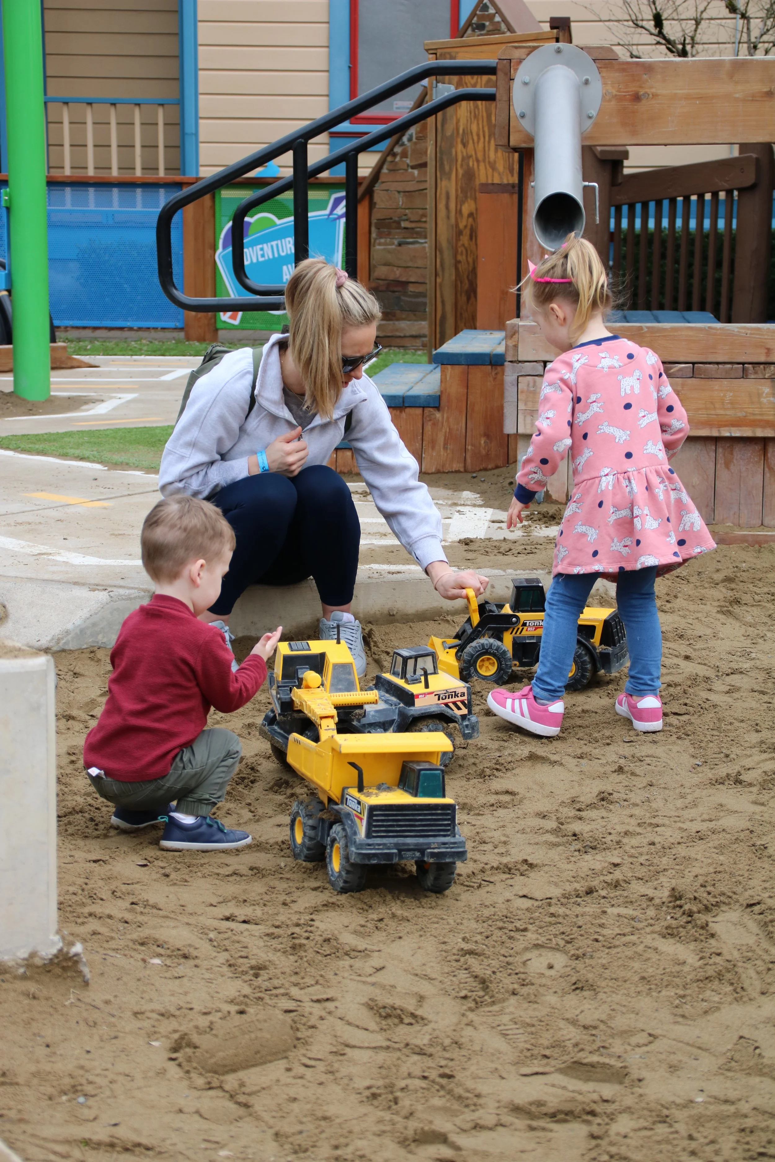A parent kneels as two toddlers, one in a red shirt and one in a pink polka-dot dress, enthusiastically play together in a large, sandy construction zone with yellow toy dump trucks.