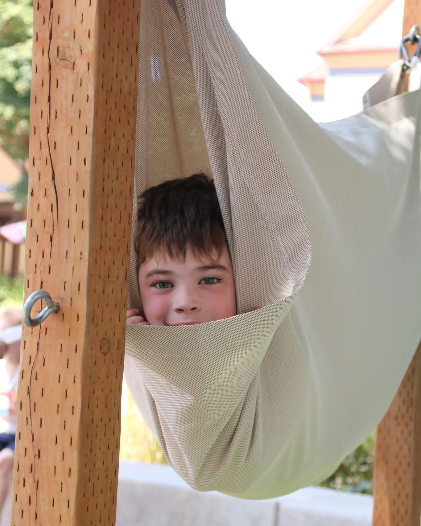 Child with brown hair lying in a beige fabric hammock hanging between wooden posts, looking at the camera with a curious expression.