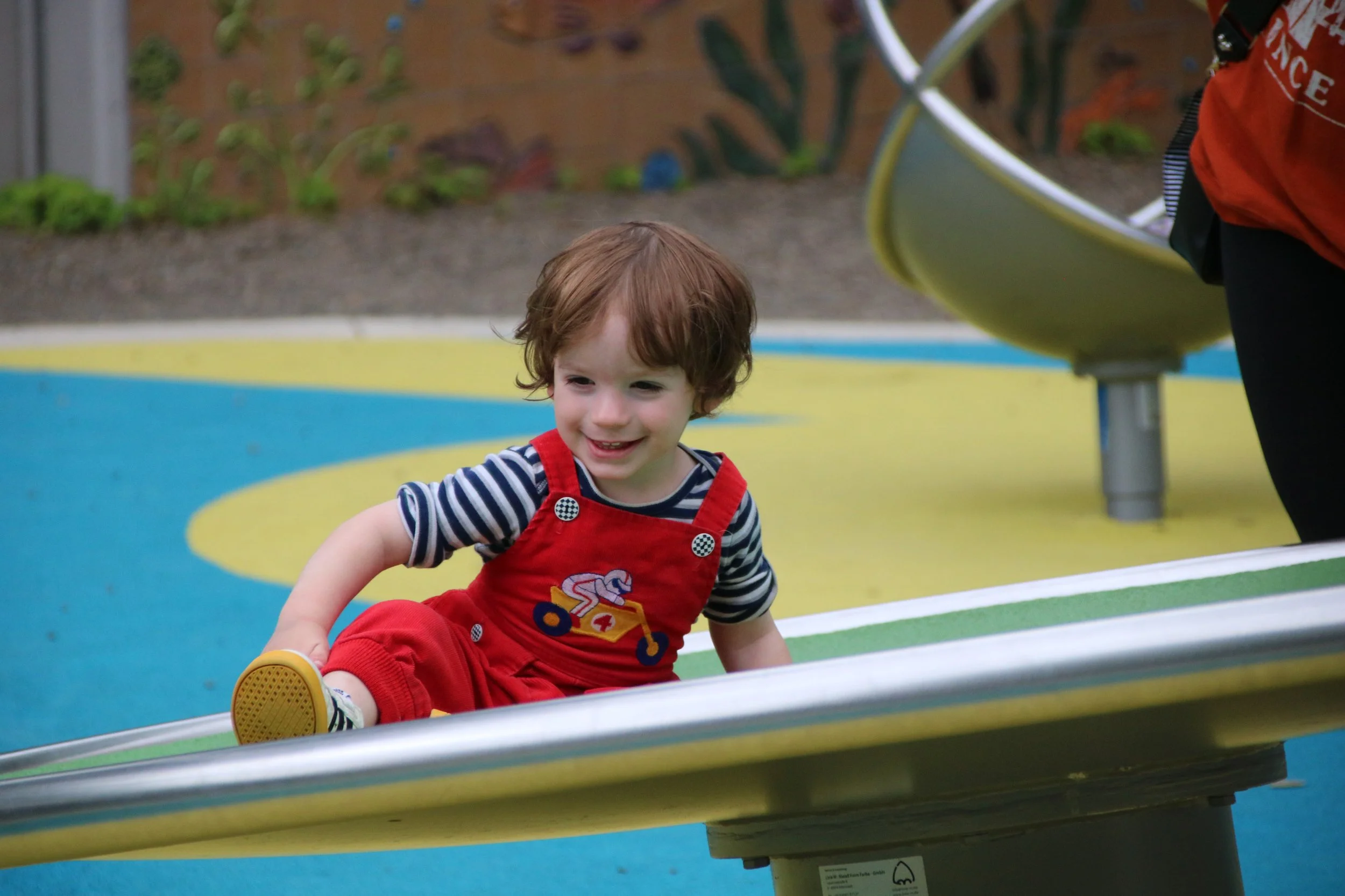 A young boy with brown curly hair smiling and sitting on a colorful playground teeter-totter, wearing red overalls with a striped shirt.