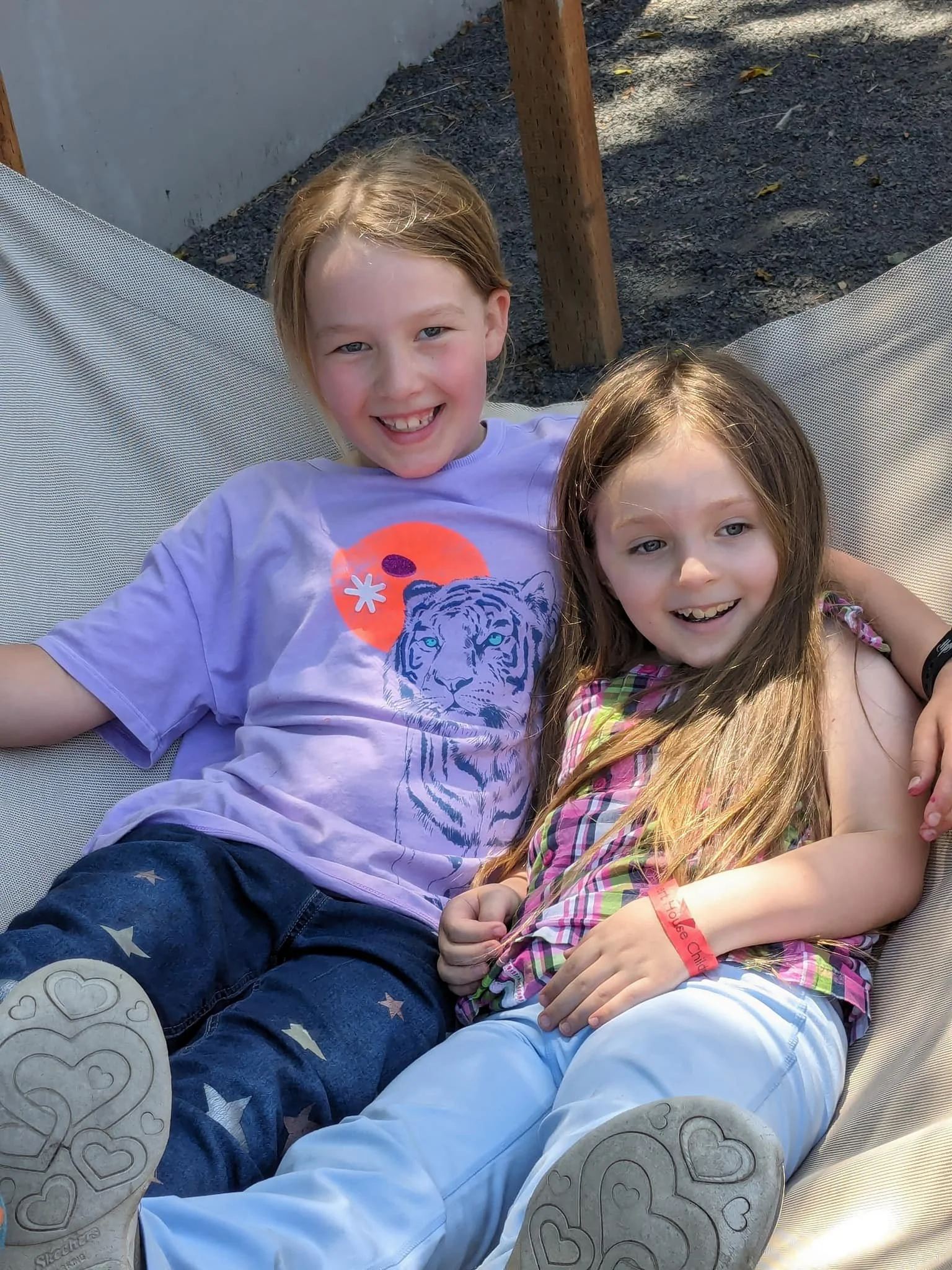 Two young girls are smiling and sitting together on a light-colored outdoor swing or seat. The girl on the left has blonde hair and is wearing a purple t-shirt with a tiger graphic and colorful design, and star-patterned pants. The girl on the right has light brown hair, a plaid top, and white pants. They are outdoors with some wooden posts and gravel visible in the background.