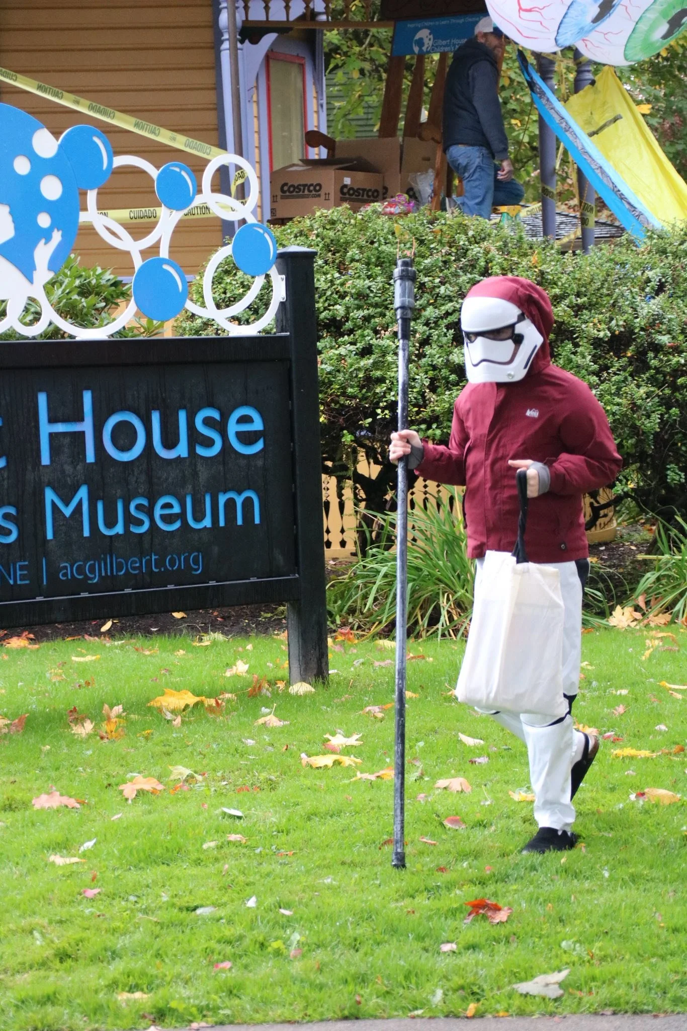 A person dressed in a Red Bull-themed costume with a Stormtrooper helmet, standing on a lawn near a sign for the Children's Museum. The person is holding a white bag and a cane. In the background, there are people, trees, and a building.