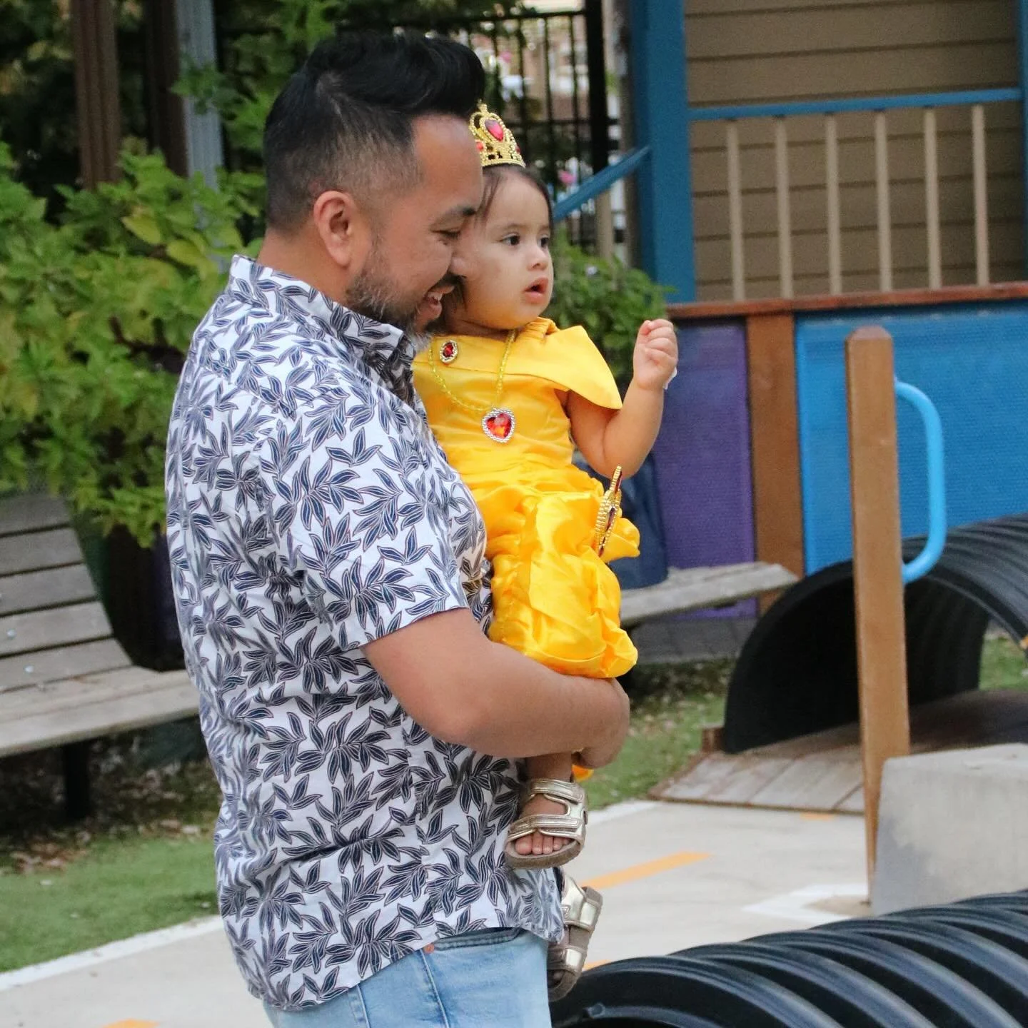 A father in a floral shirt carries his daughter, dressed as Belle, through the museum’s outdoor play area.