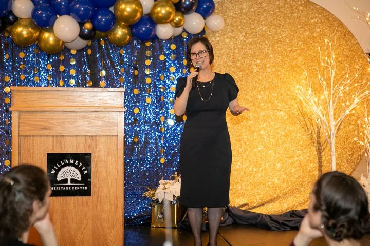A woman in a black dress speaking into a microphone at a decorated event with blue, white, and gold balloons and a glittery gold backdrop at WIlliamette Heritage Center.