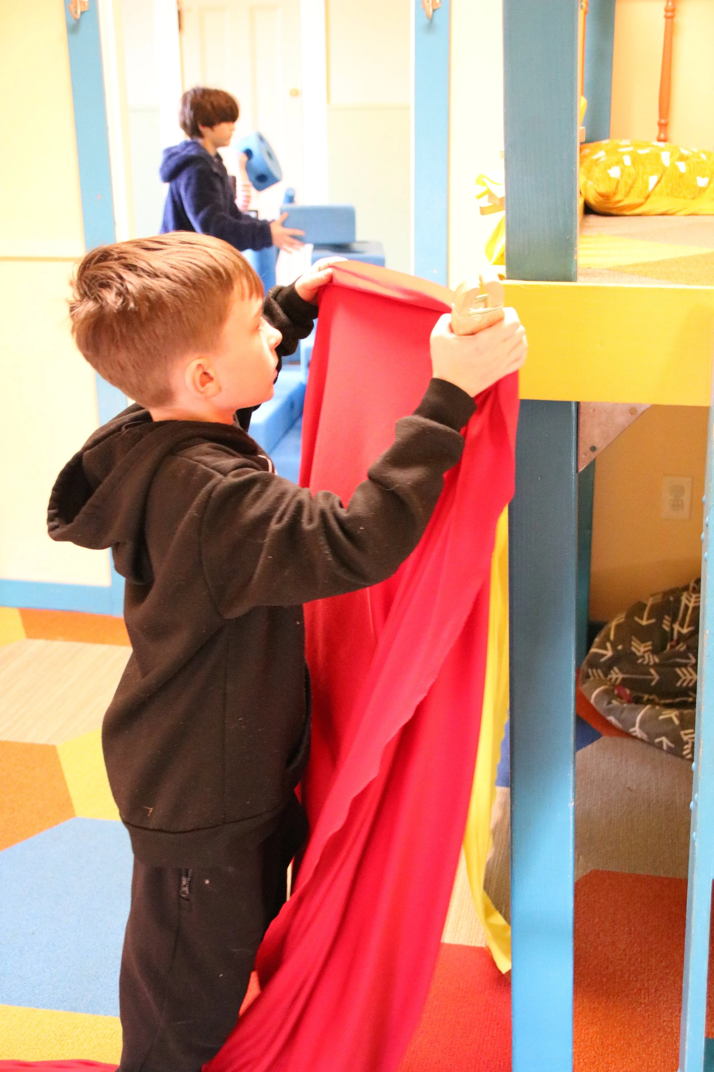 A young boy with short brown hair, wearing a black hoodie, is pulling back a red fabric curtain in a brightly colored indoor play area. In the background, another child with dark hair and a blue hoodie is holding a blue toy. The play space has yellow, blue, and orange walls, with a bed or cushion visible in the upper right corner.