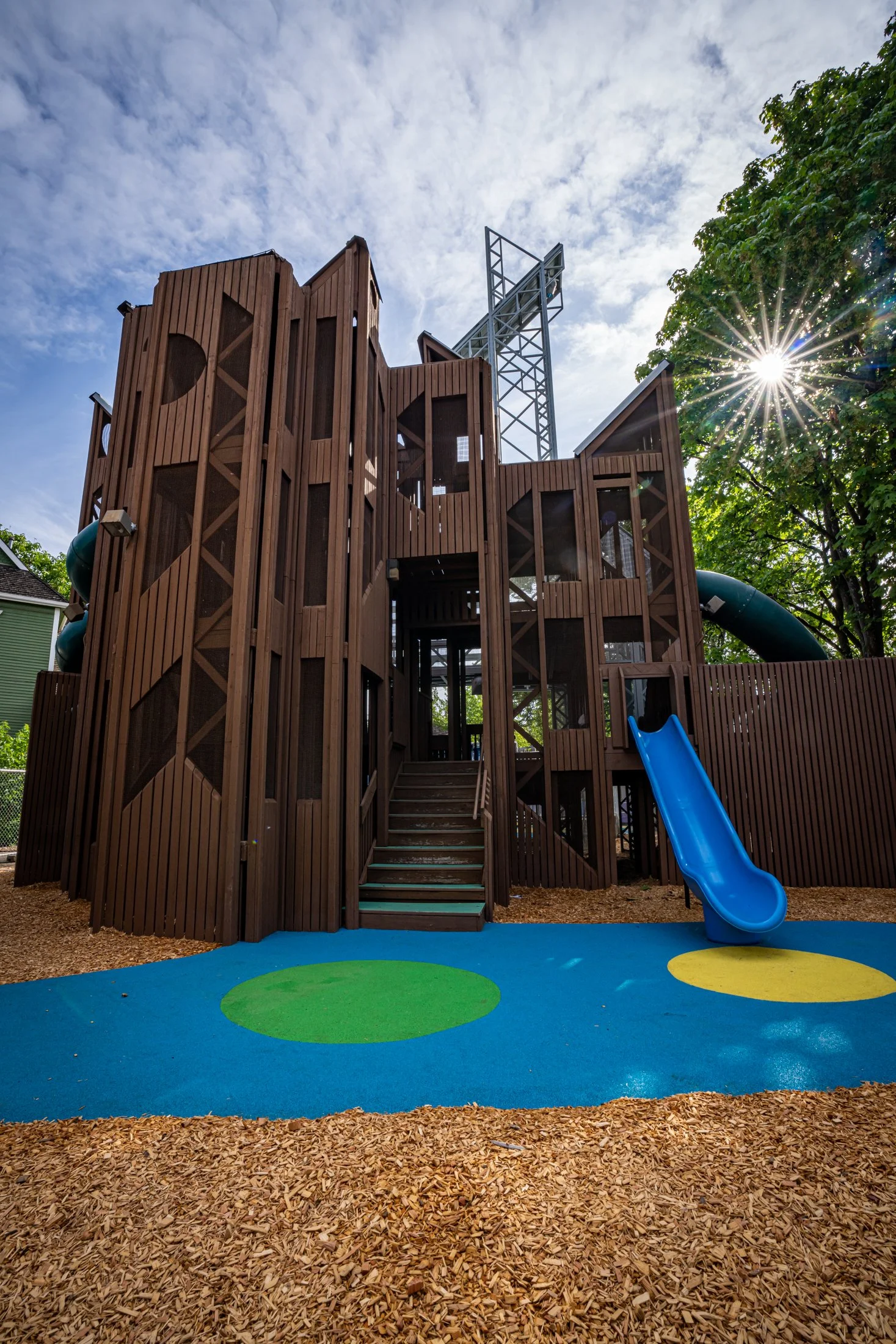 Towering, multi-level dark brown wooden play structure featuring stairs and a blue slide, designed for climbing, building, and active exploration.