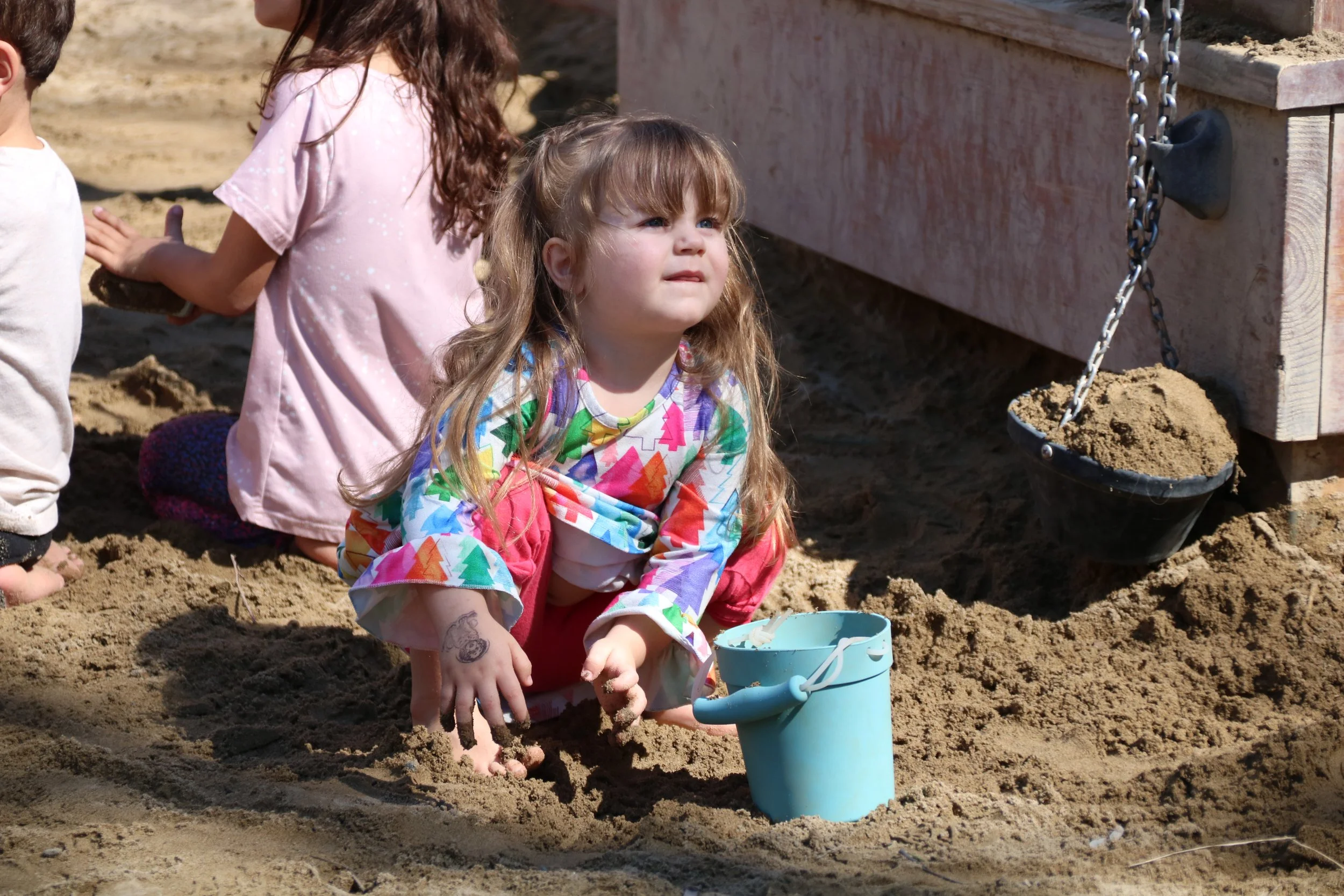 A young girl with long, wavy blonde hair sitting in the sand, holding a blue toy bucket, wearing a colorful, patterned long-sleeve shirt and pink pants. Other children are partially visible around her, and a sandbox structure with a chain and a bucket filled with sand is in the background.