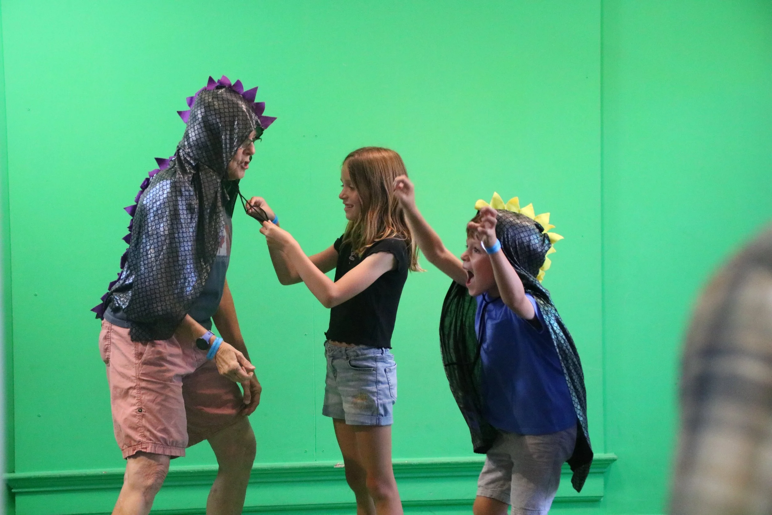 Two children dressed as dinosaurs playfully interacting with an adult in front of a green screen. One child is wearing a black and yellow dinosaur costume, and the other is in a black and purple dinosaur costume, while the adult is holding a microphone.