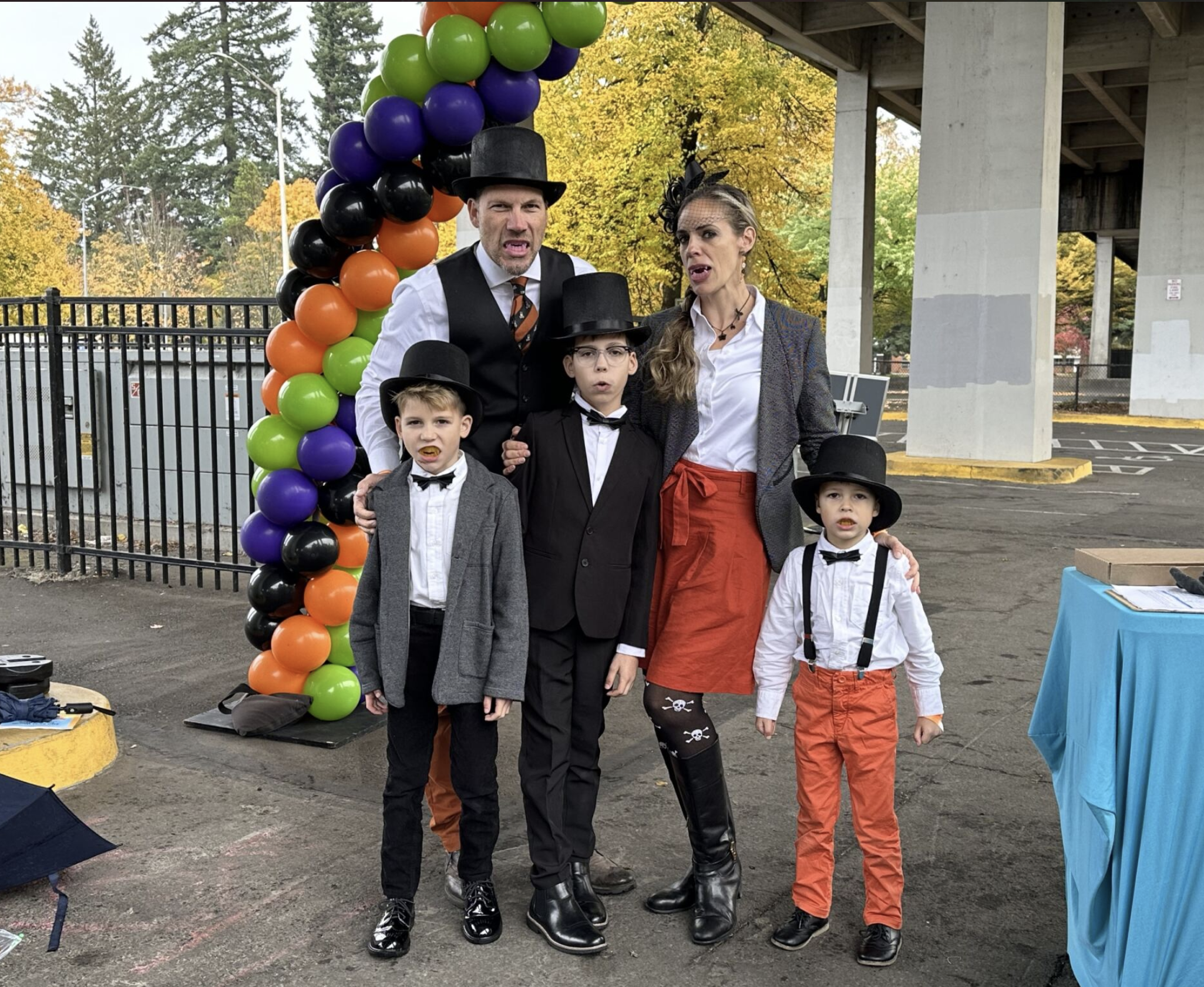 Family dressed in vintage circus costumes standing under decorated balloon arch outdoors with trees in the background.