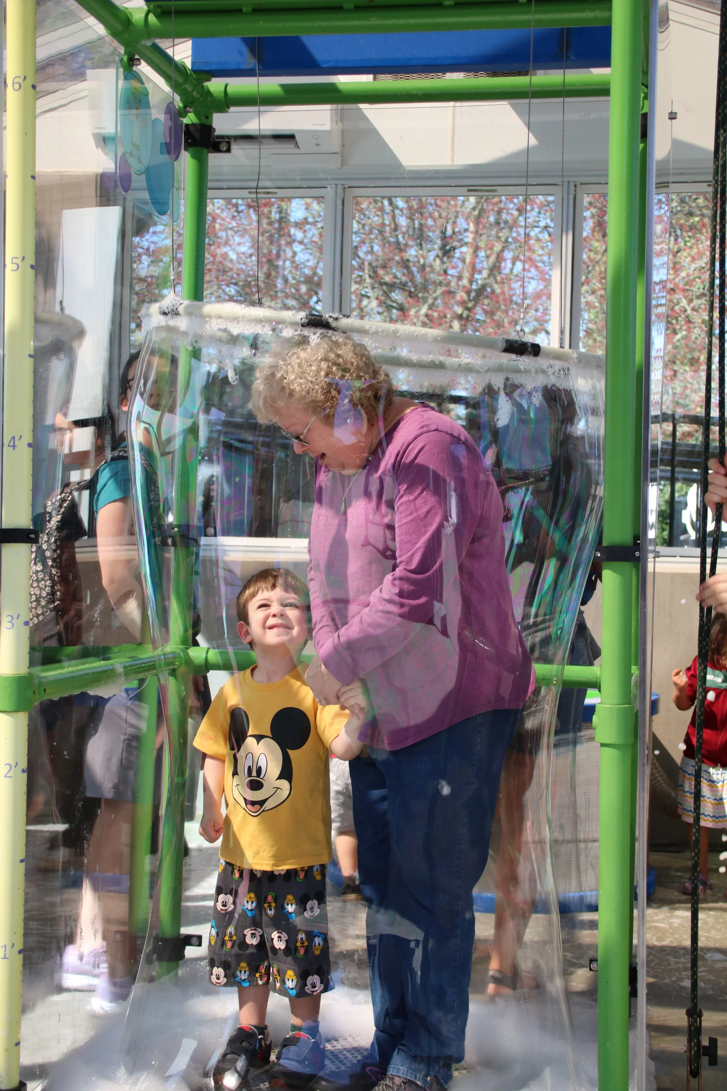 A woman and a young boy standing inside a large, transparent, soap-filled bubble on a sunny day. The boy is wearing a yellow Mickey Mouse shirt and shorts with Mickey Mouse characters. The woman is wearing a purple jacket. There are several people and trees visible in the background.