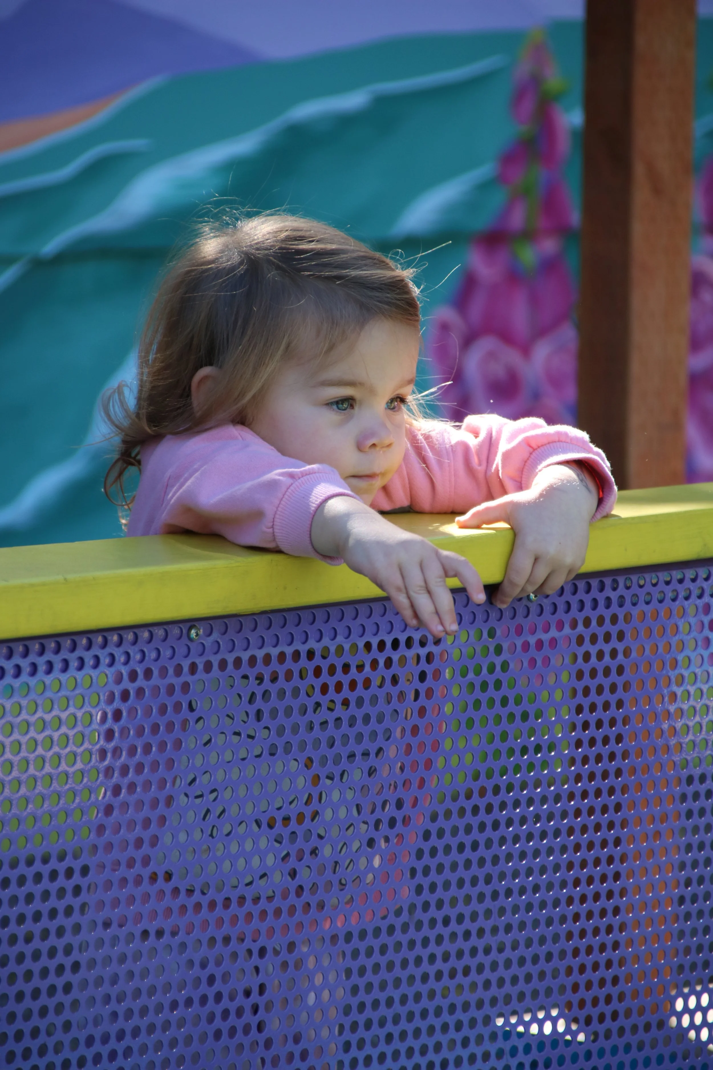 A young girl with brown hair and blue eyes leans over a purple and yellow playground barrier, looking pensively into the distance on a sunny day.