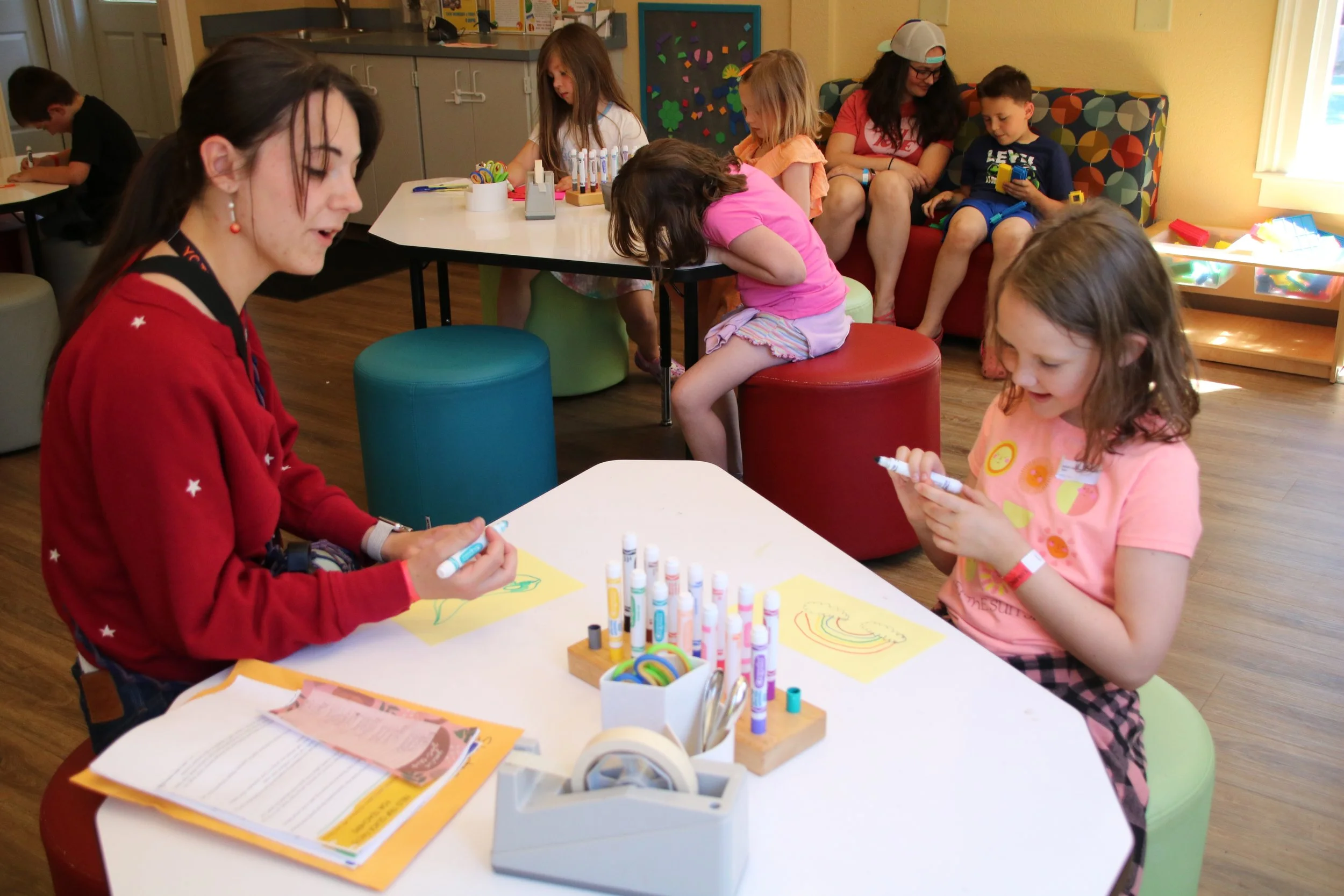 A group of children and an adult engaging in arts and crafts activities at a table in a colorful room with yellow walls, colorful stools, and art supplies.