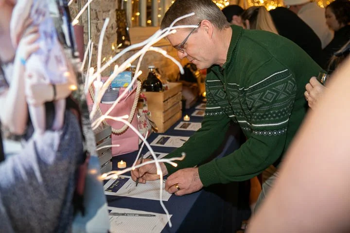 A man in a green sweater signing or writing on a table covered with a navy cloth at a public event, with other people around him and decorative lights in the background.