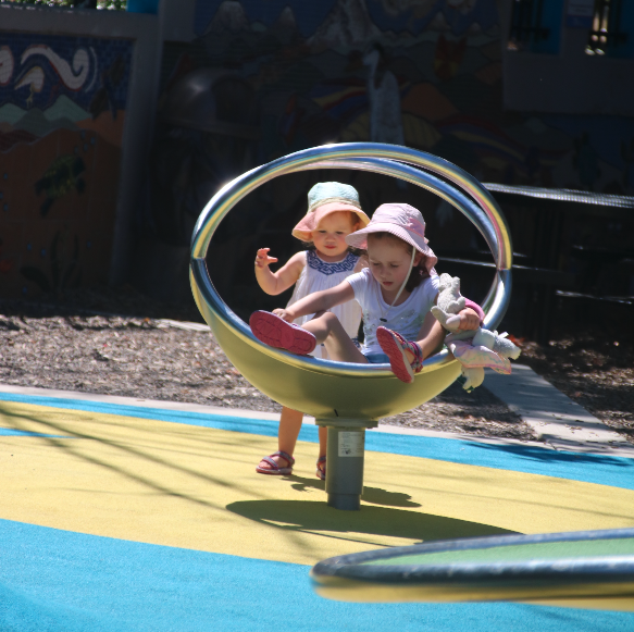 Two young children playing on a spinning amusement ride outdoors, wearing hats and summer clothes.