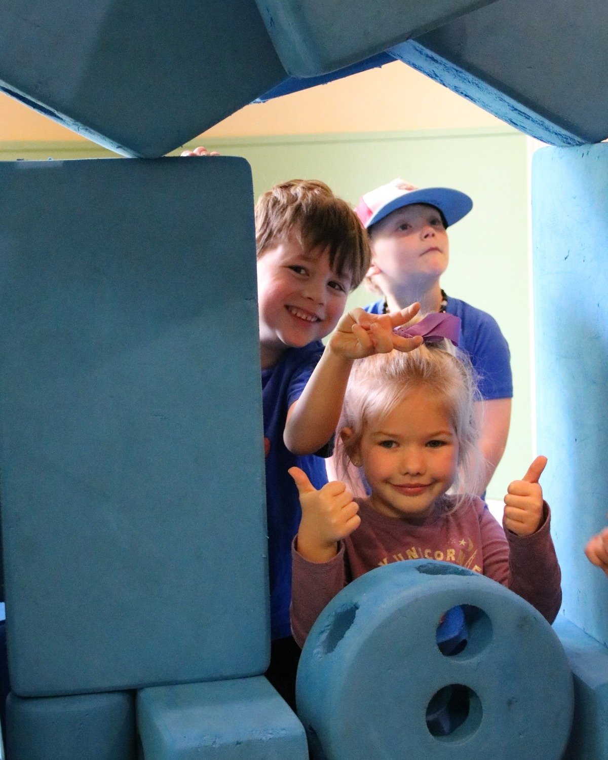 Three children play behind large blue foam blocks, smiling and giving thumbs up, inside a play area with pastel-colored walls.