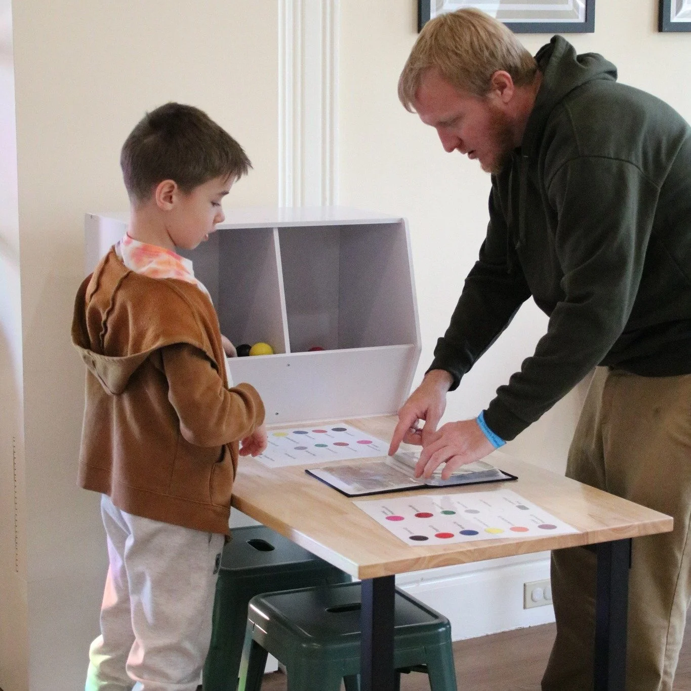 A man and a boy standing at a small table, looking at colored dots on paper and a digital device. The man is pointing at the device, and the boy is observing.