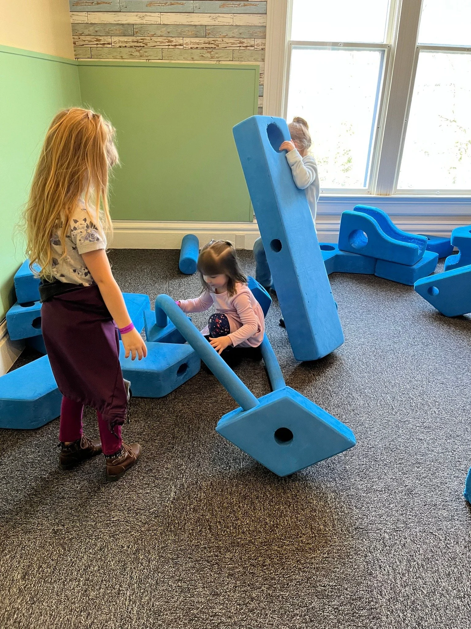 Three young children playing with large blue foam building blocks in an indoor play area near a window.