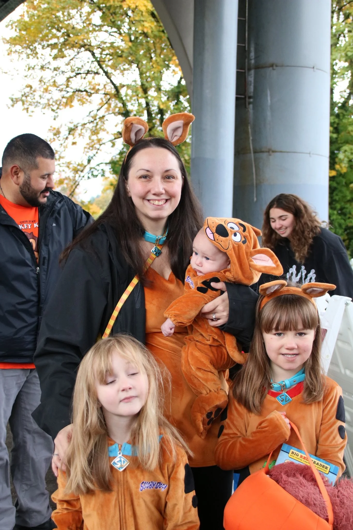 A group of people dressed in Scooby-Doo costumes, including a woman and three children, outdoors during daytime with trees and a bridge in the background.
