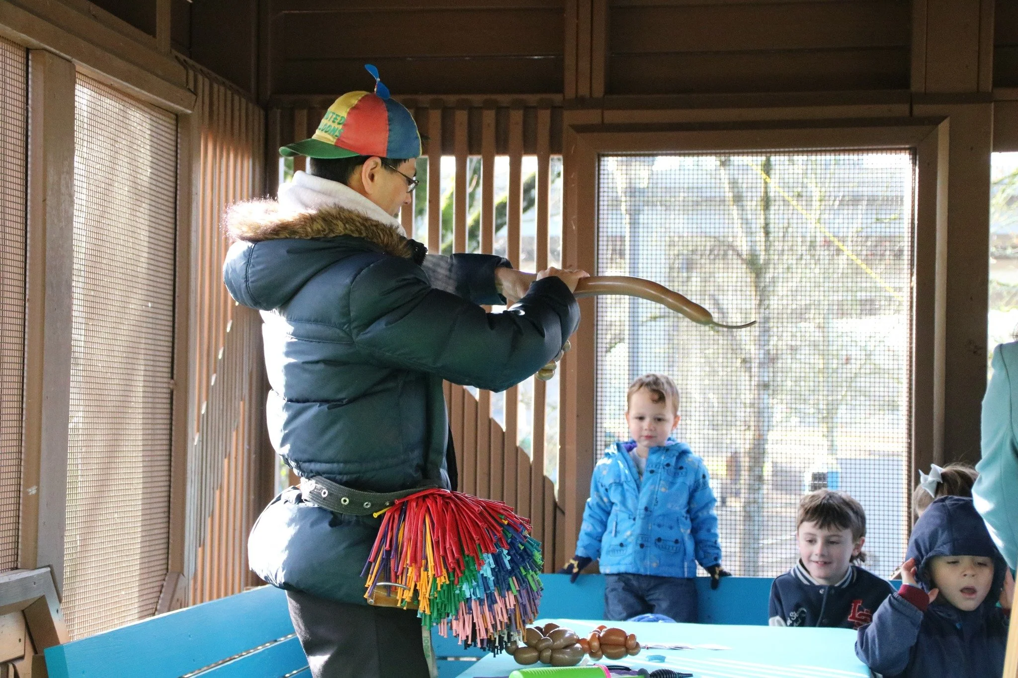 A person wearing a colorful cap, glasses, and a thick winter coat with a fur-lined hood is holding a long eel inside a wooden enclosure with children watching nearby.