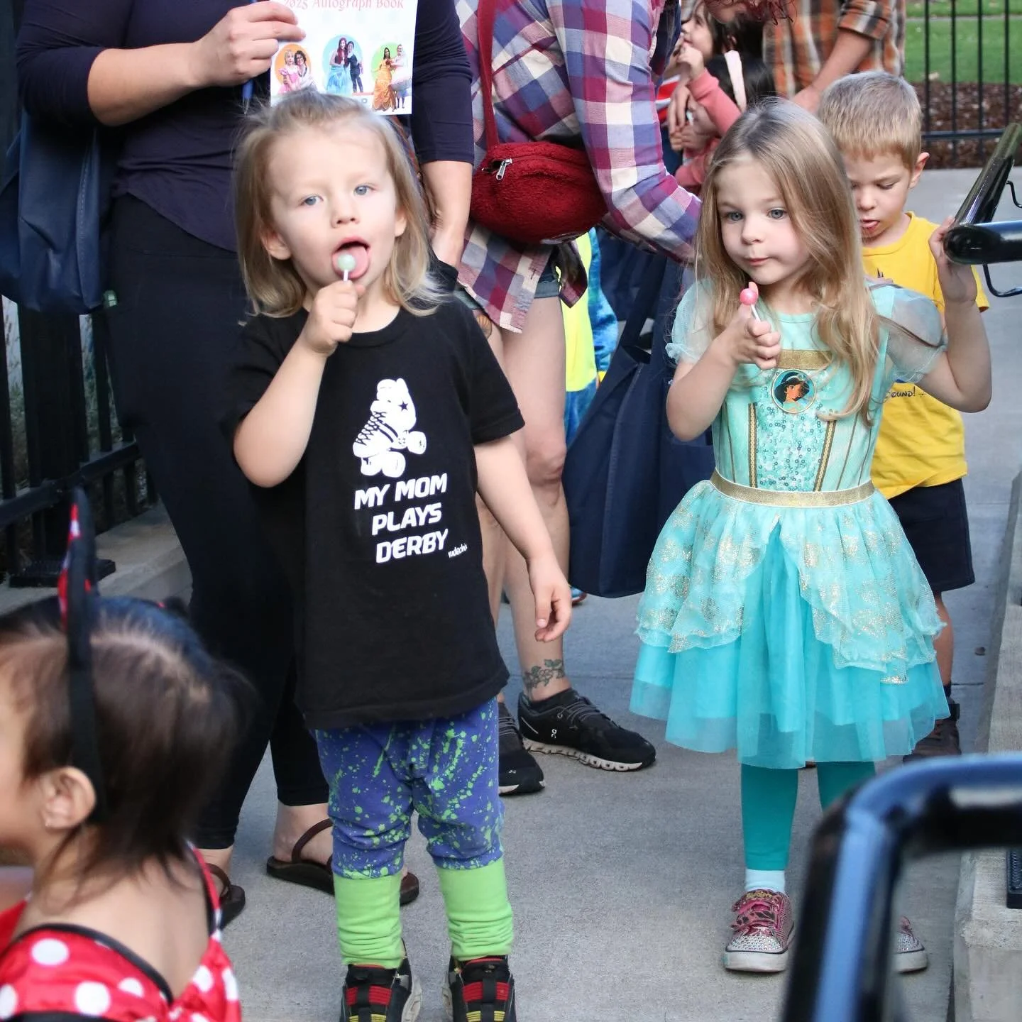 Two young girls and several children in the background, one girl dressed as a princess and the other in a black t-shirt with a dog graphic, standing outdoors on a sidewalk, holding ice cream treats.