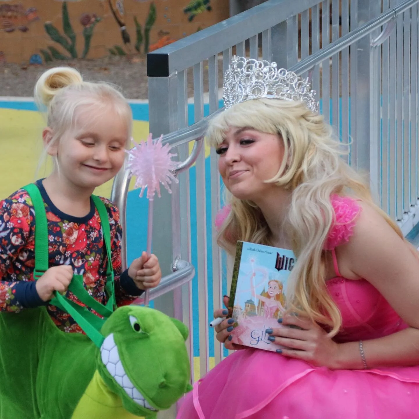 A princess character in pink smiles at a young girl holding a green dinosaur plush toy at a museum celebration.