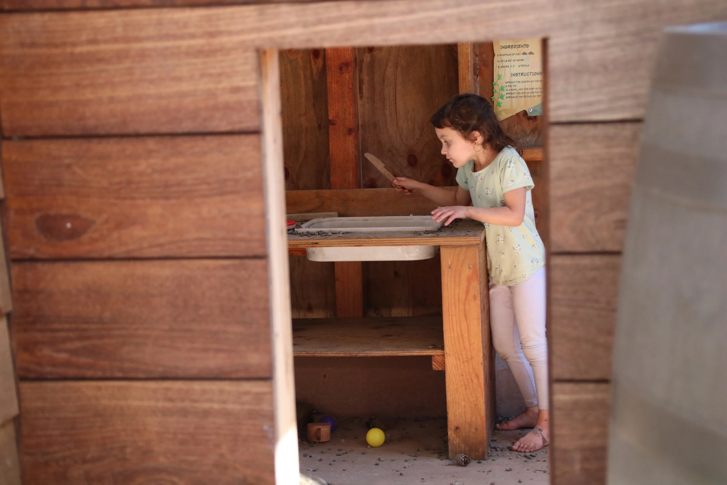 A young girl inside a wooden playhouse, playing with sand and using a small shovel or scoop.