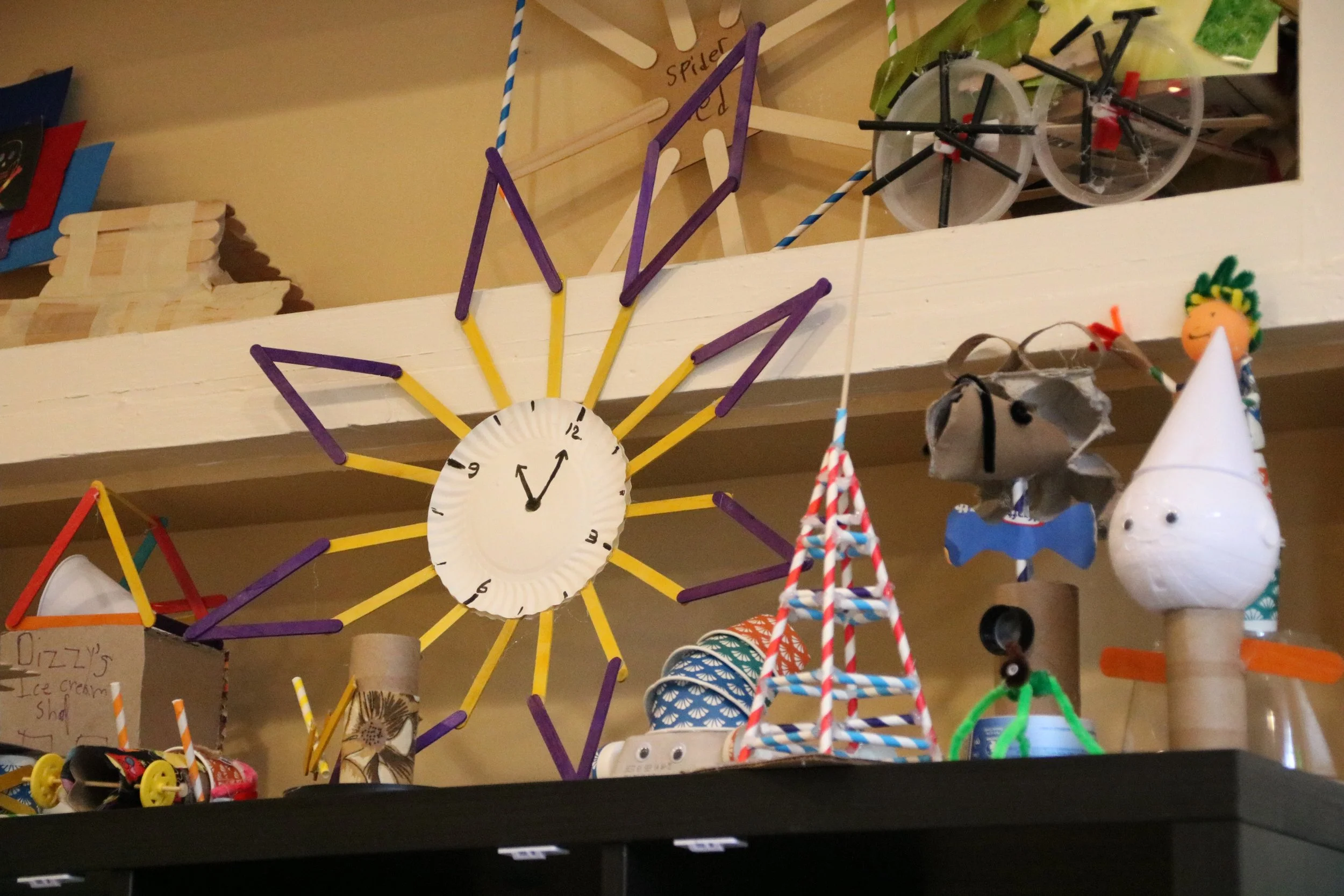 A display of colorful handmade crafts and clocks on a shelf, including a sun-shaped clock with purple and yellow sticks, a small model bicycle, and various other decorative items.